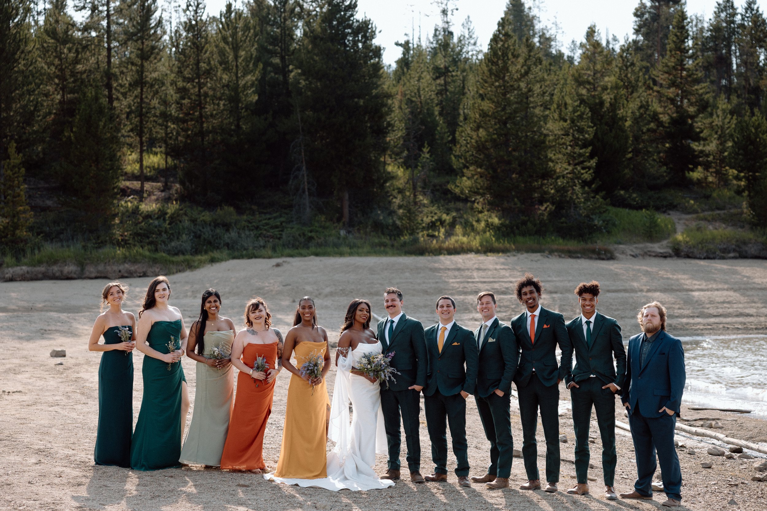 A wedding party standing on a sandy beach with a forest background, featuring bridesmaids in colorful dresses and groomsmen in suits, with a bride in a white gown and a groom in a dark suit.
