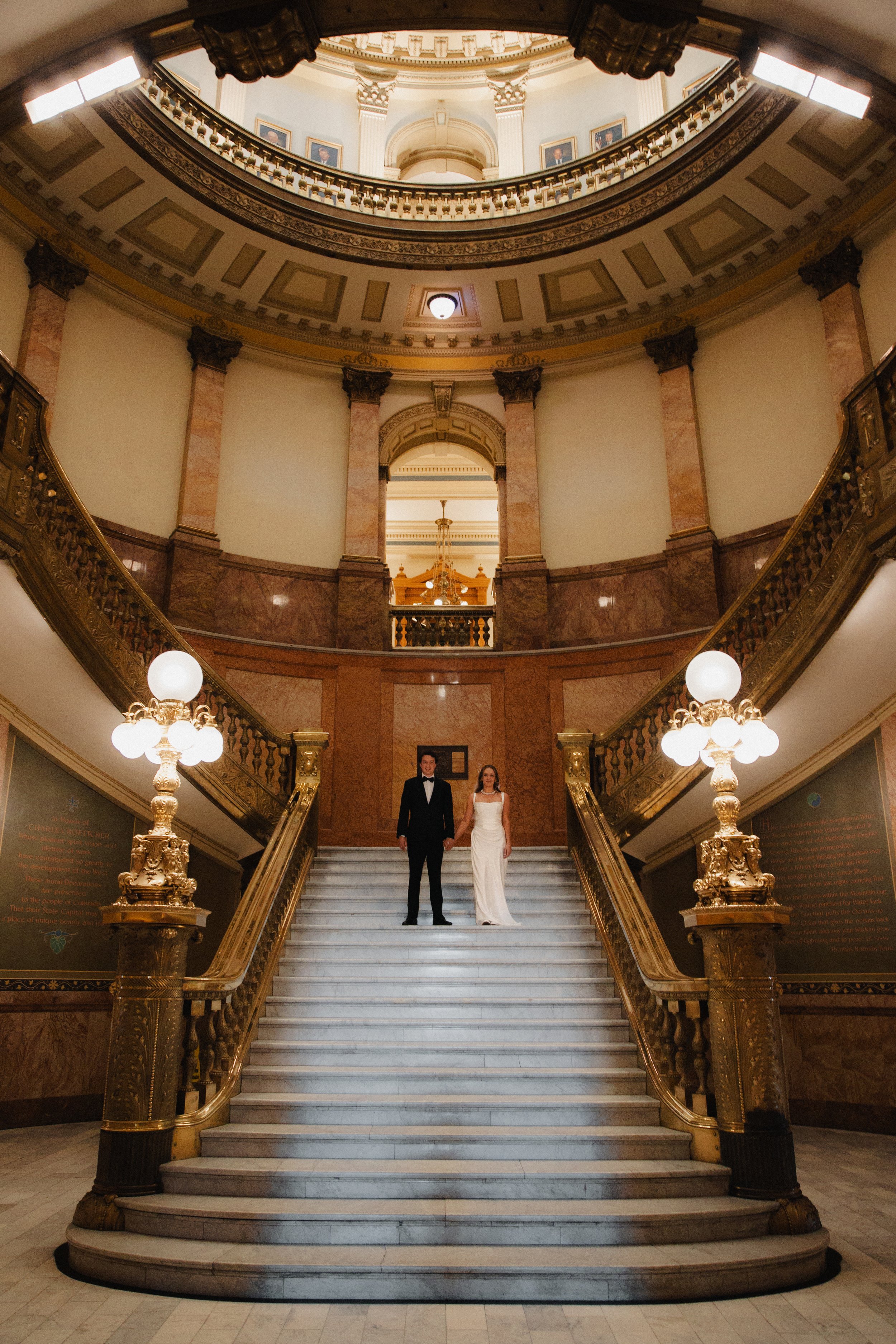 A couple dressed in wedding attire standing on a grand staircase inside a historic, ornate building.