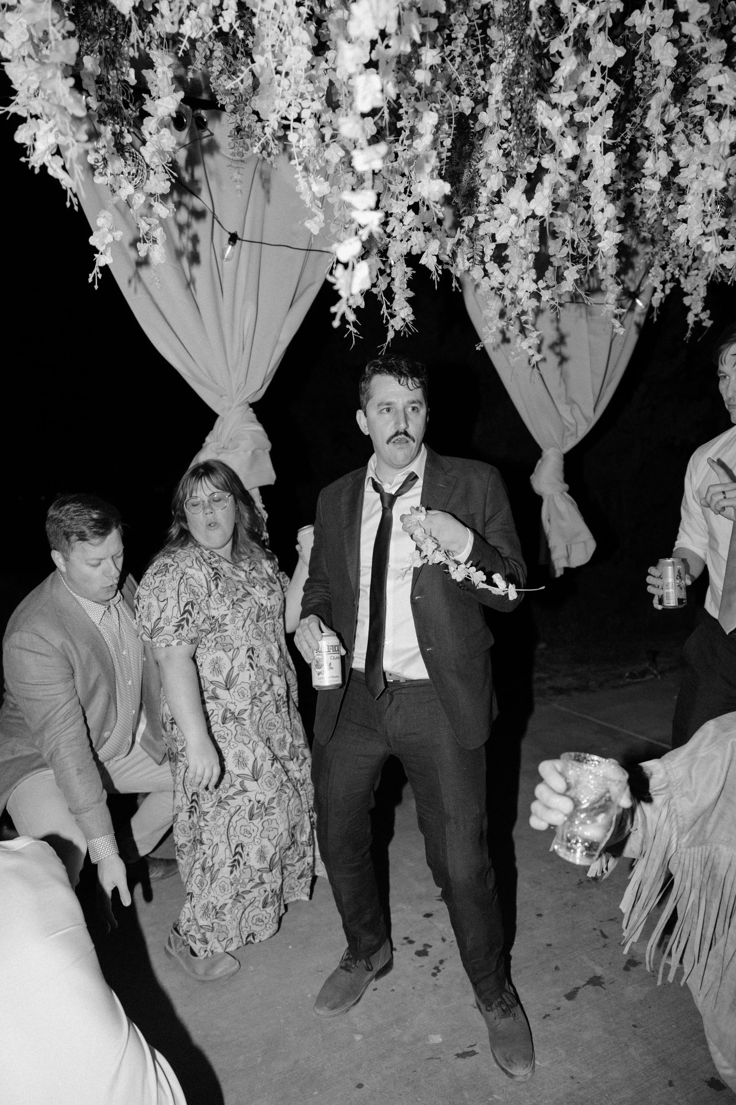 A black and white photo of a group of people dancing at a party, with a decorated canopy of flowers above them. The man in the center is holding a drink and wearing a suit with a loosened tie and a lei around his neck.