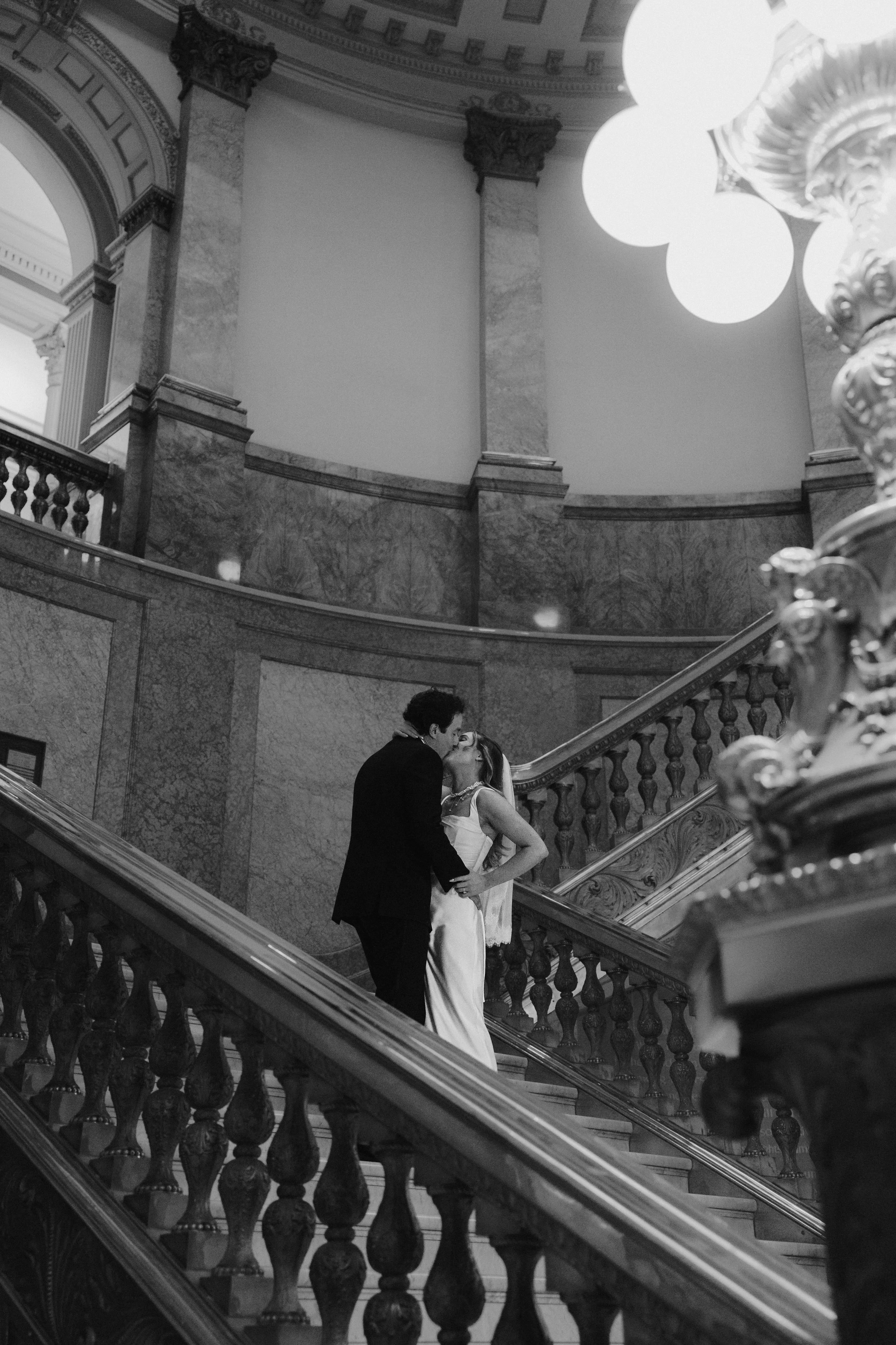 A black and white photo of a couple kissing on a grand marble staircase inside a historic building with ornate architectural details and a large light fixture overhead.
