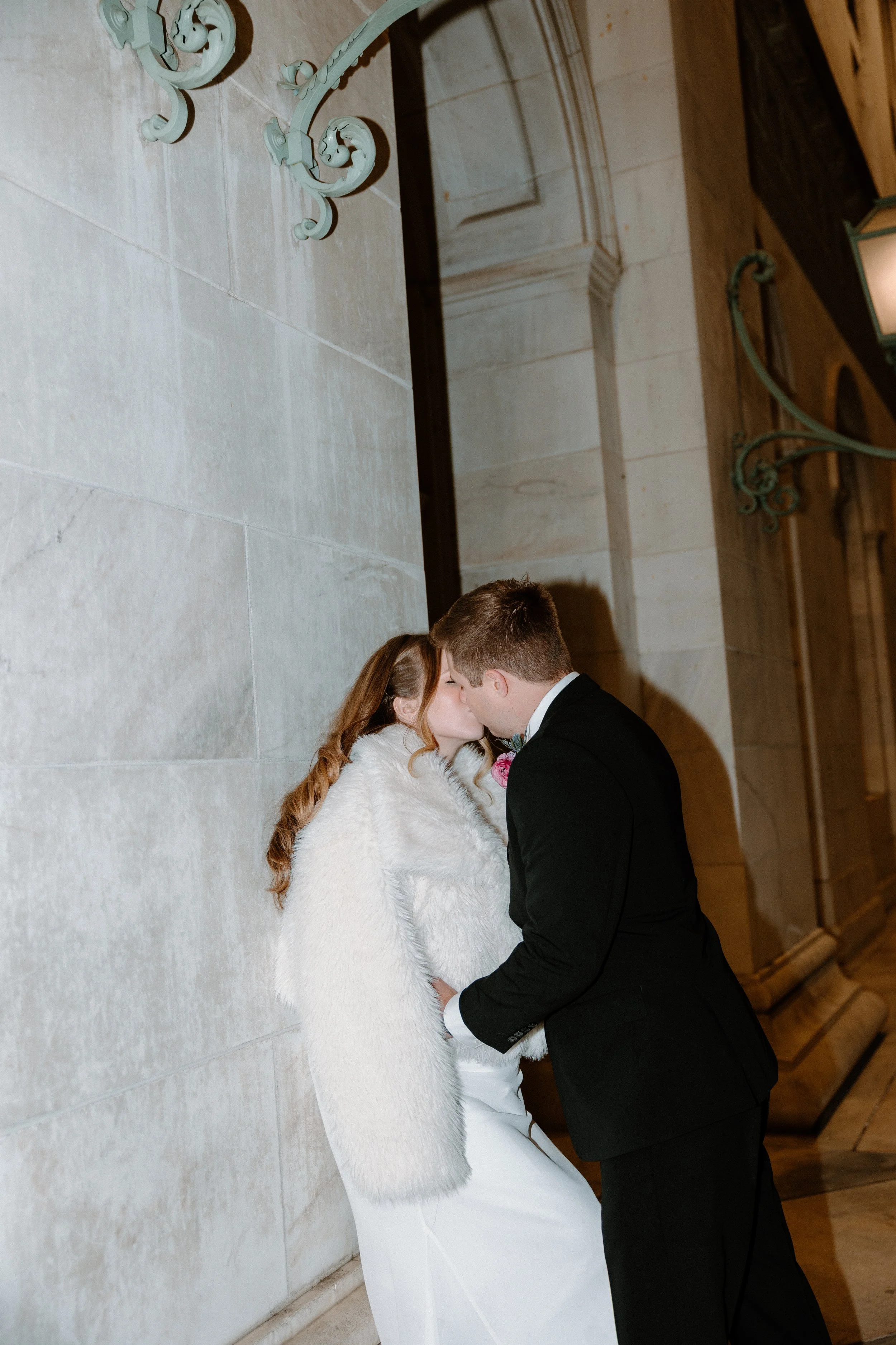A couple in formal attire sharing a kiss, the woman wearing a white dress and fur coat, leaning against a stone wall with ornate metalwork, in an indoor setting with elegant architecture.