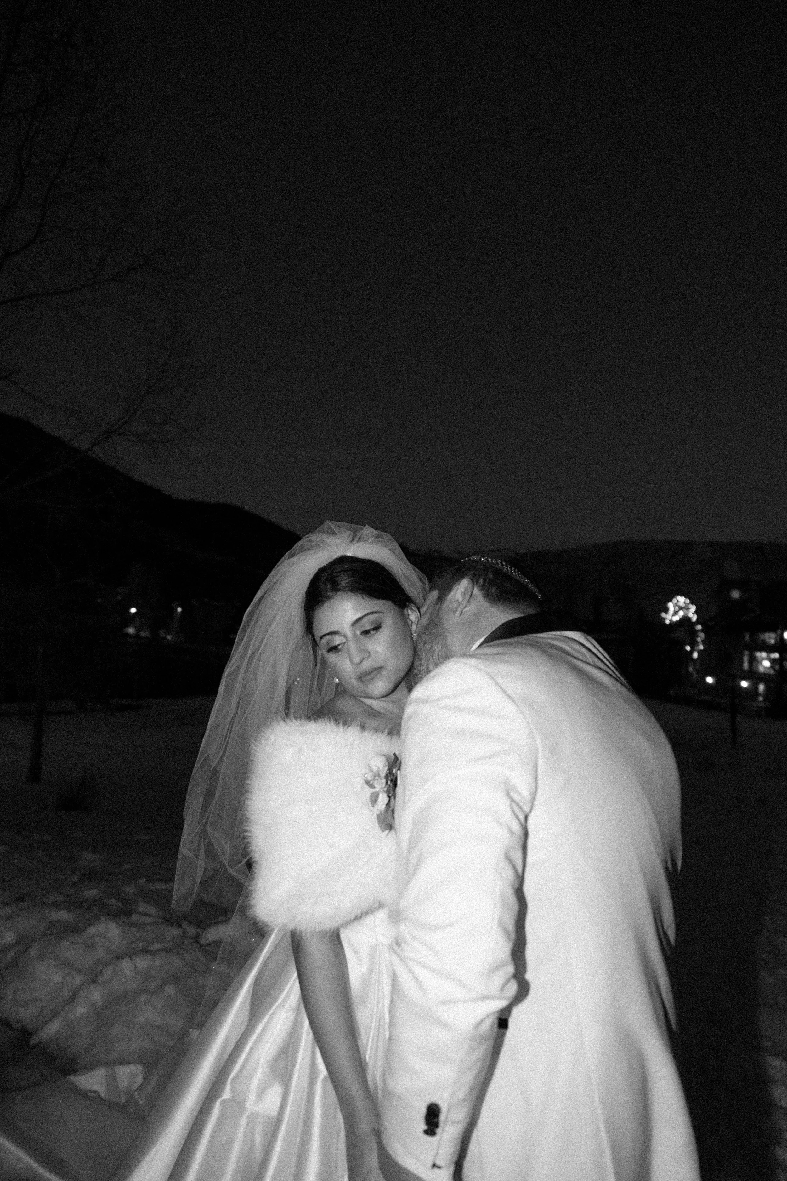 A black and white photo of a bride in a wedding gown and veil, and a groom in a tuxedo, sharing an intimate moment outdoors at night.