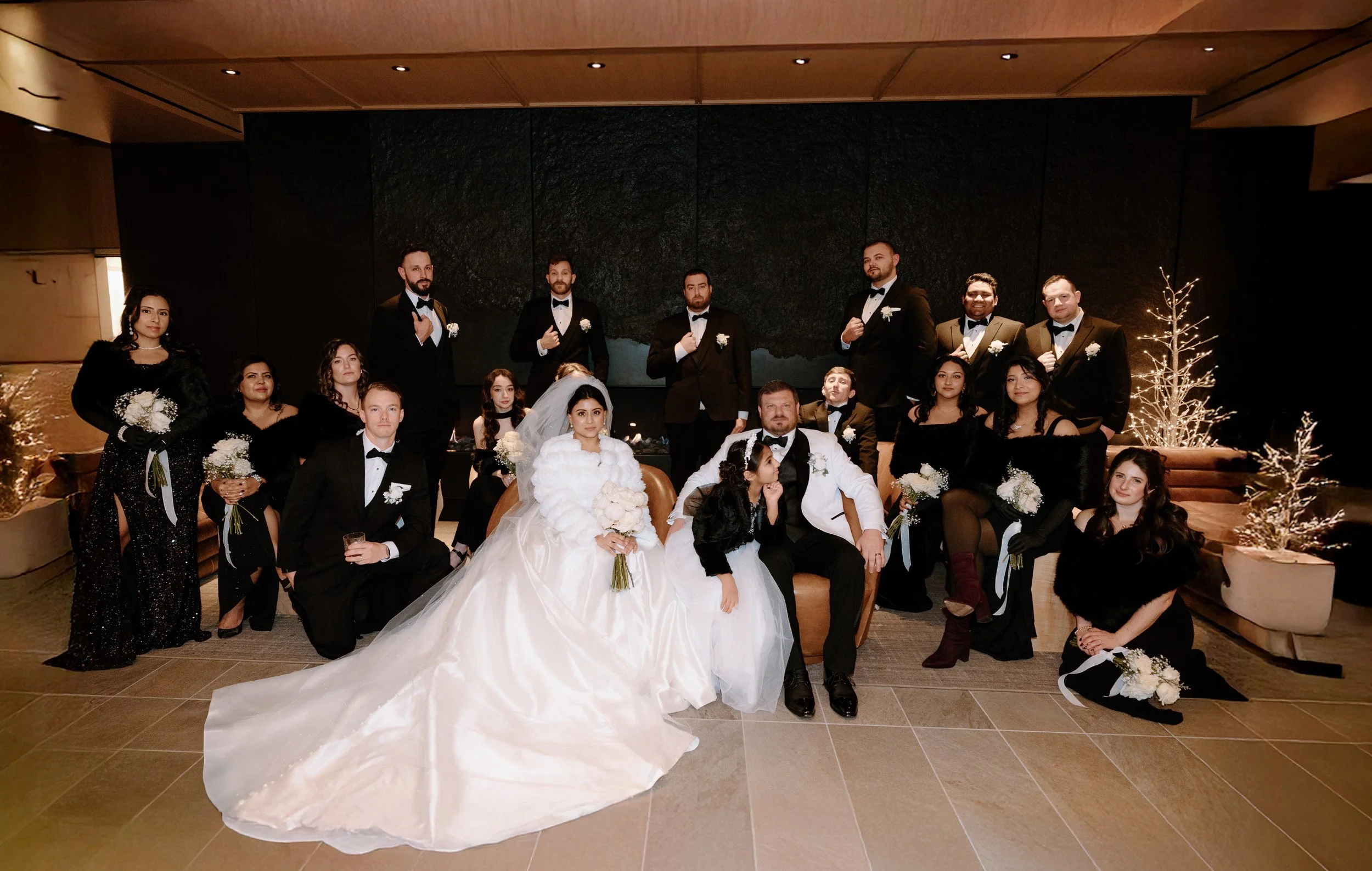 Wedding party with the bride in a white gown and groom in a tuxedo, surrounded by bridesmaids and groomsmen in formal attire, posed indoors with holiday decorations.