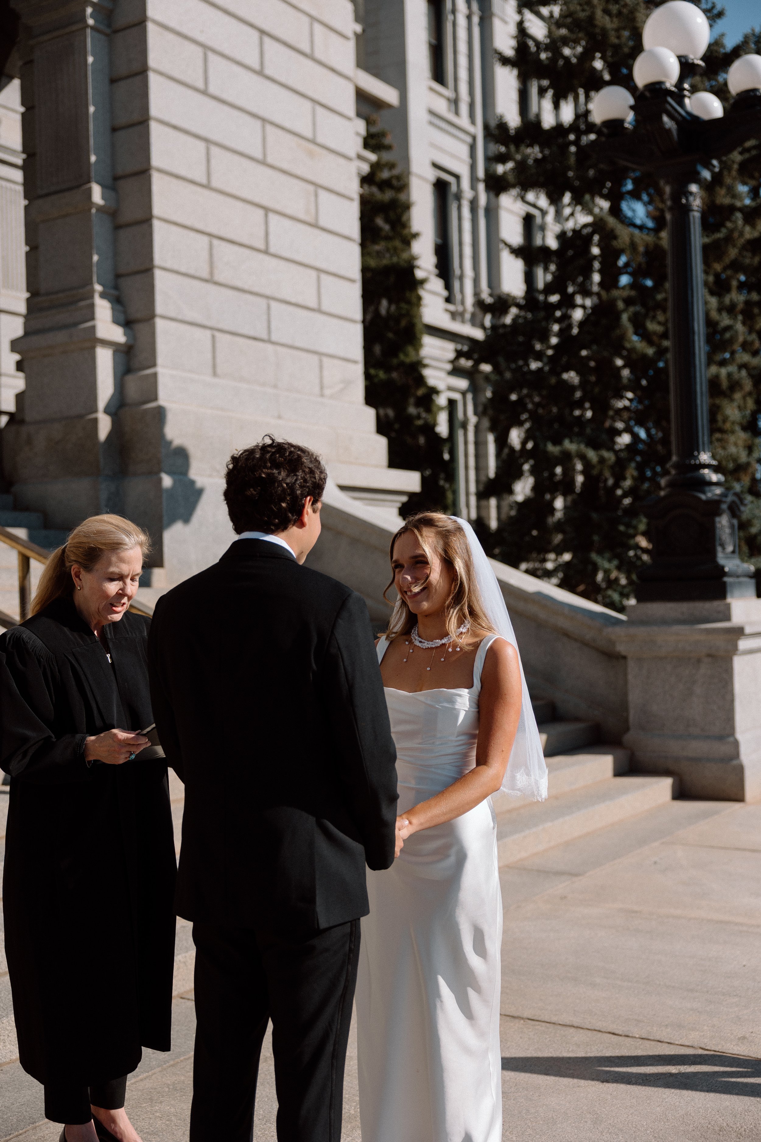 A bride and groom holding hands during their wedding ceremony outside a courthouse, with a woman officiant reading vows.