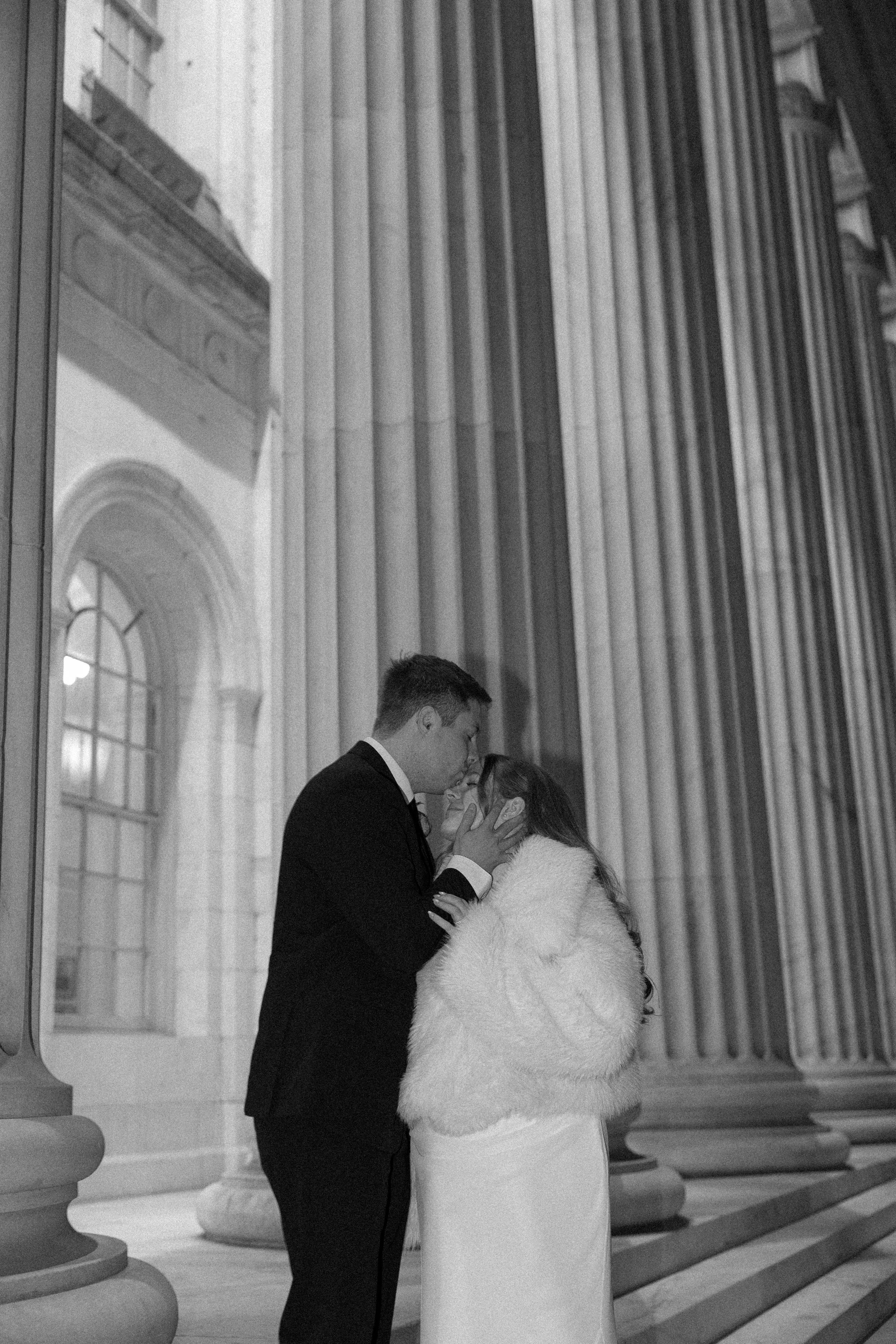 A black and white photo of a couple in formal attire having a romantic moment in front of large classical columns at a historical building, likely during a wedding or special event.