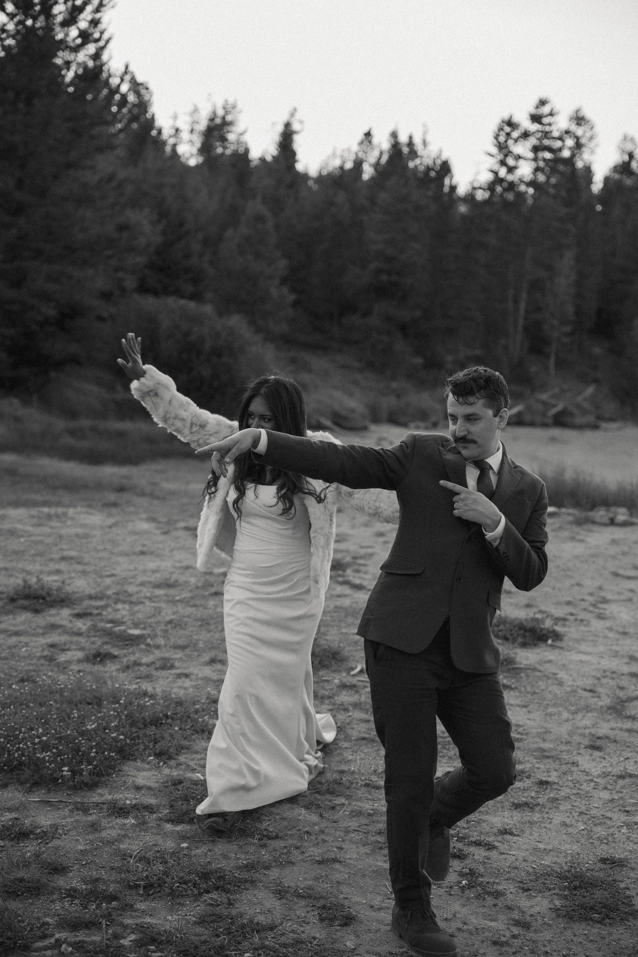A black and white photo of a man and woman dancing outdoors on a dirt path near forested area. The man is wearing a suit and the woman is in a gown with a fur jacket, both appearing to be enjoying themselves.