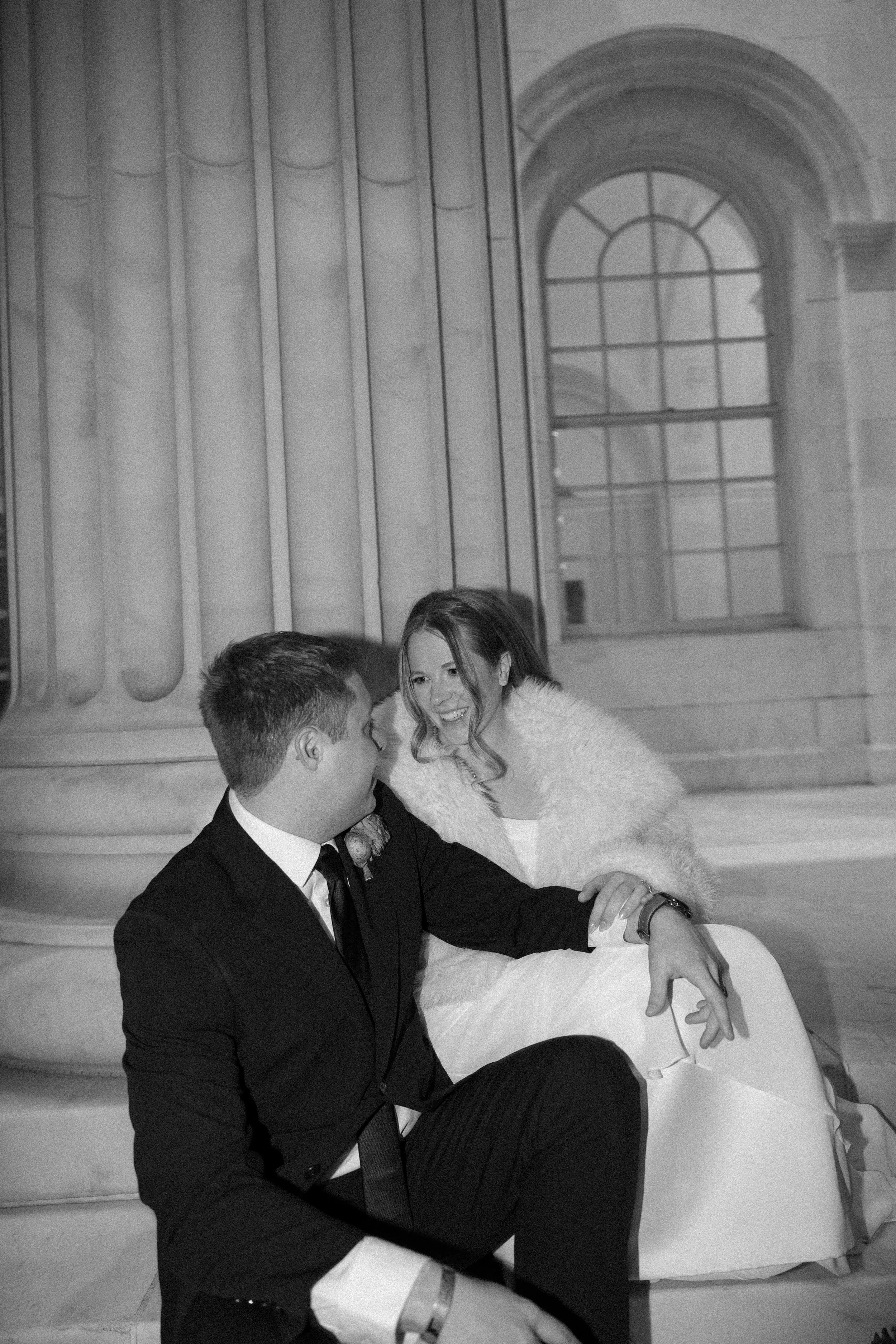 A black and white photo of a bride and groom sitting together on steps outside a building with columns and a large window, smiling and talking.
