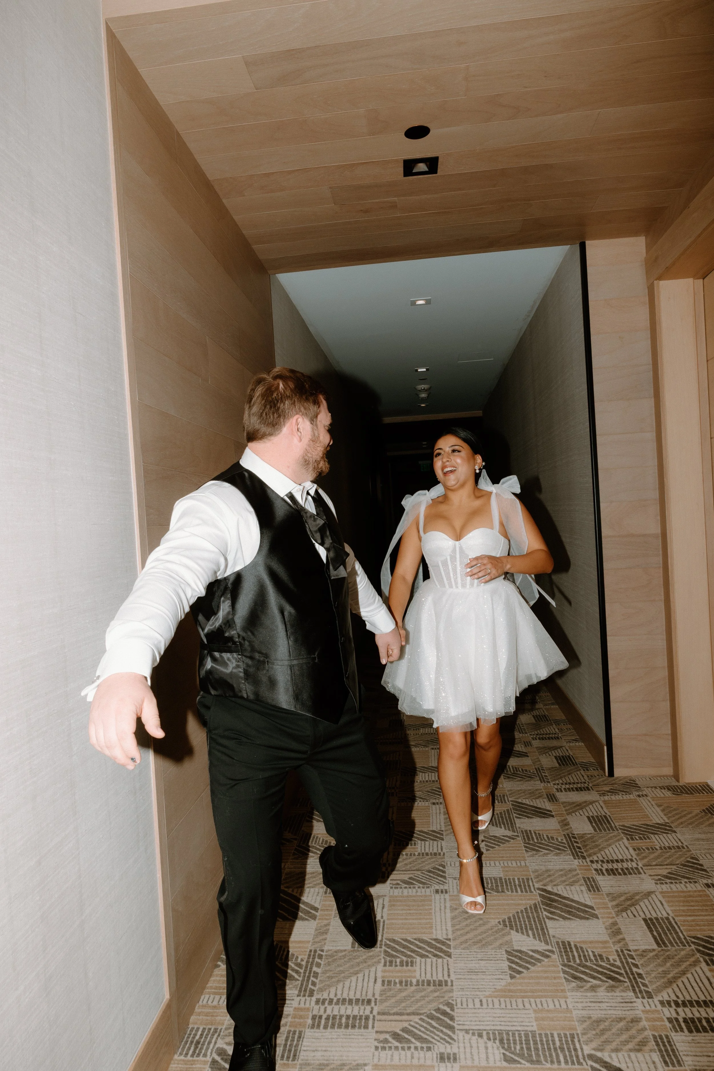 A couple dressed in formal attire, holding hands and smiling, walking down a hallway during a celebration.