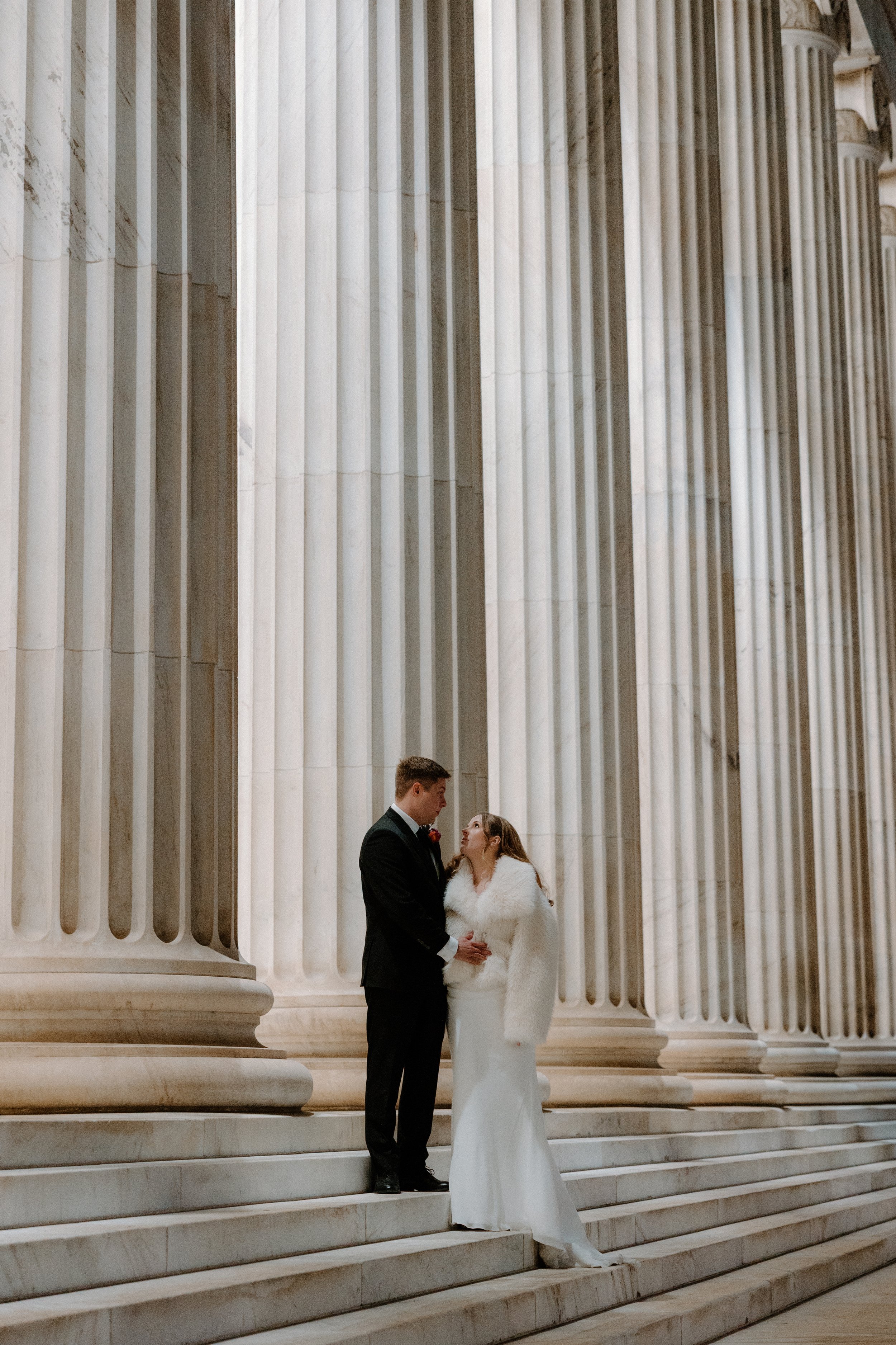 A bride and groom standing on marble steps outside a building with large columns, holding hands and looking at each other.