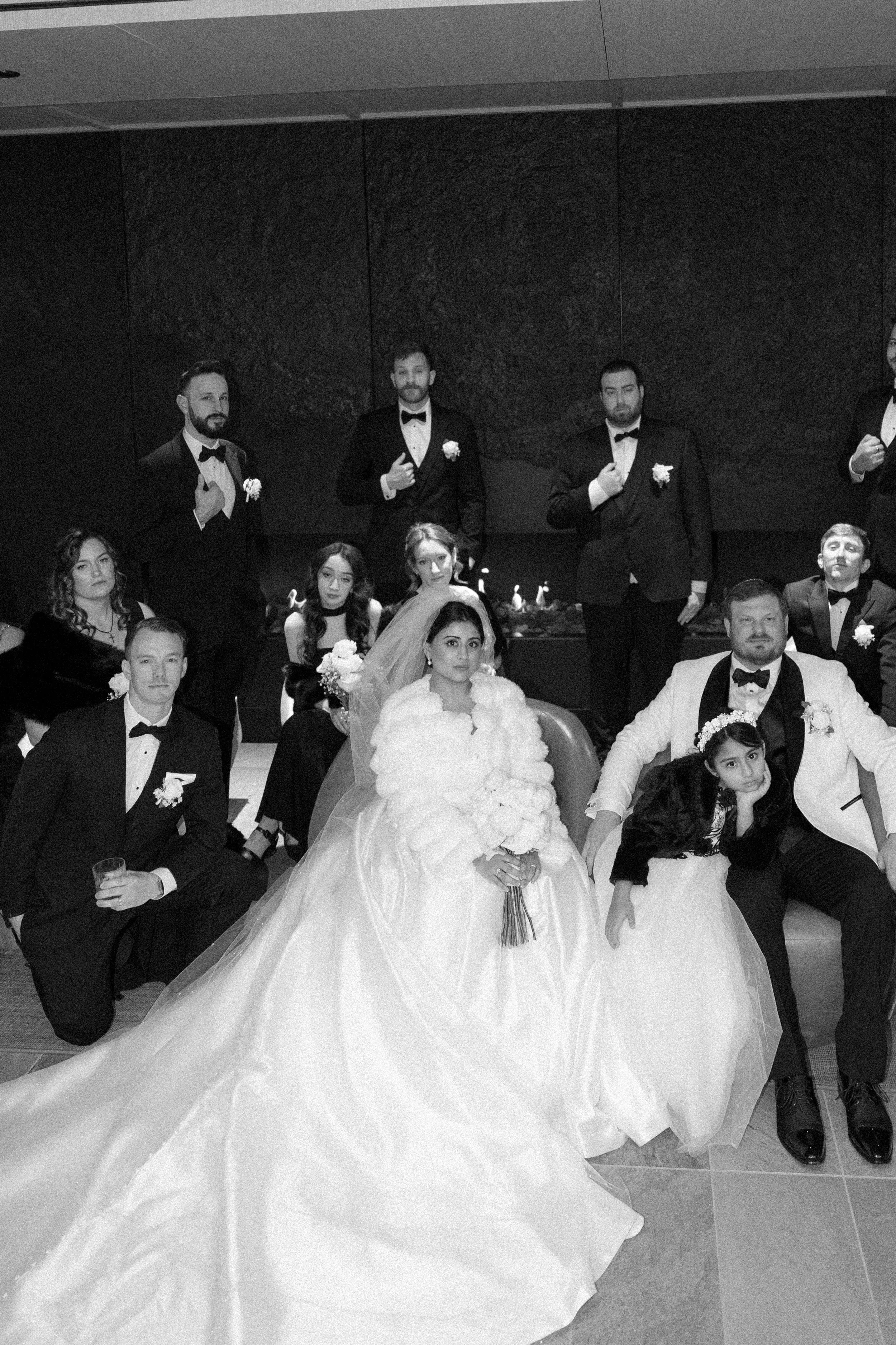 Group photo of wedding party with bride and groom in wedding attire, surrounded by friends and family in formal wear, in an indoor venue.