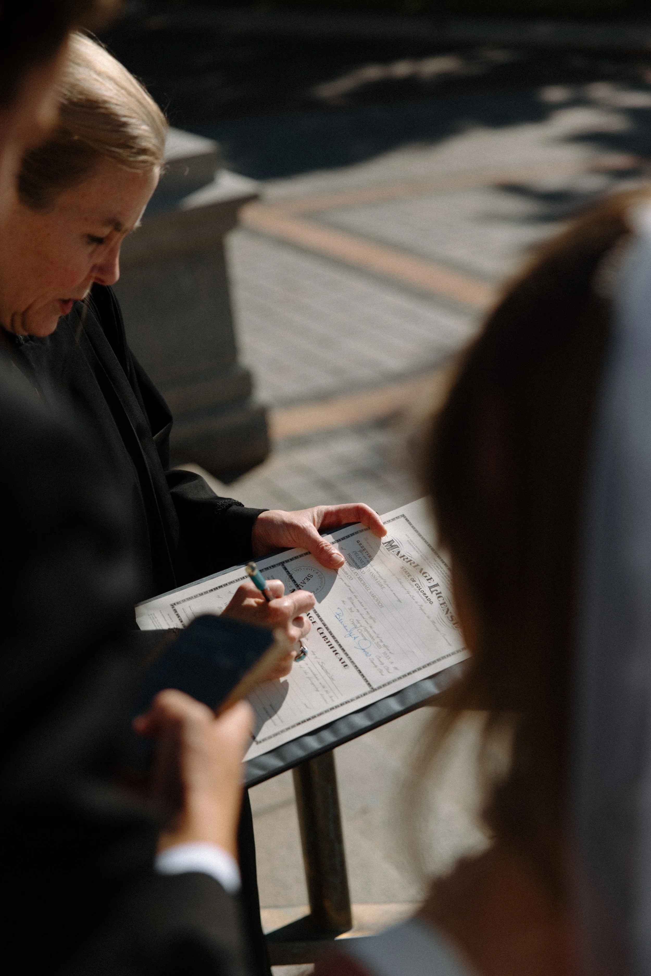 A woman signing a marriage license outdoors on a sunny day, with a person holding a phone nearby.