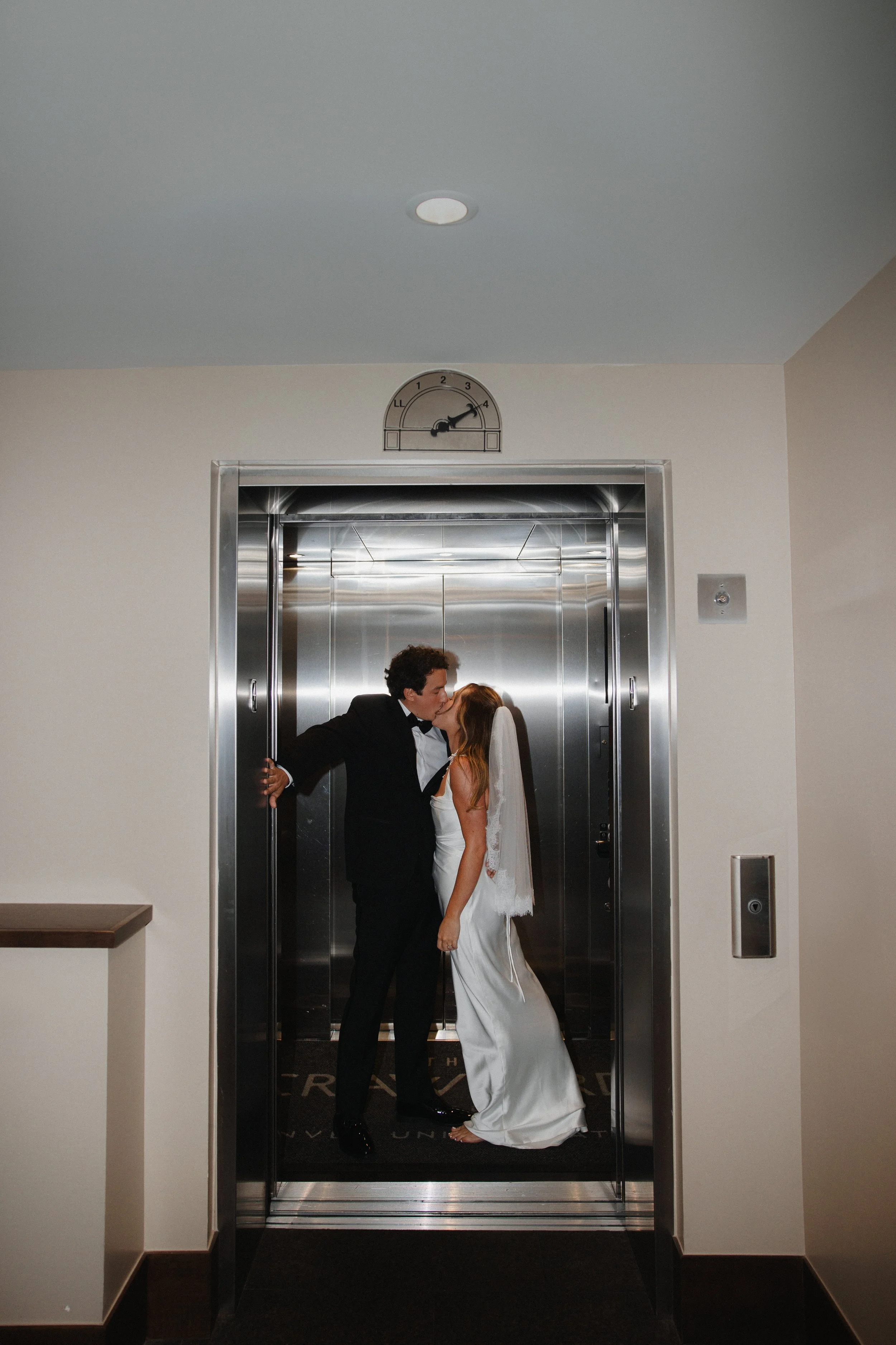 A bride and groom sharing a kiss in an elevator, with a wedding dress and tuxedo, in a modern building.