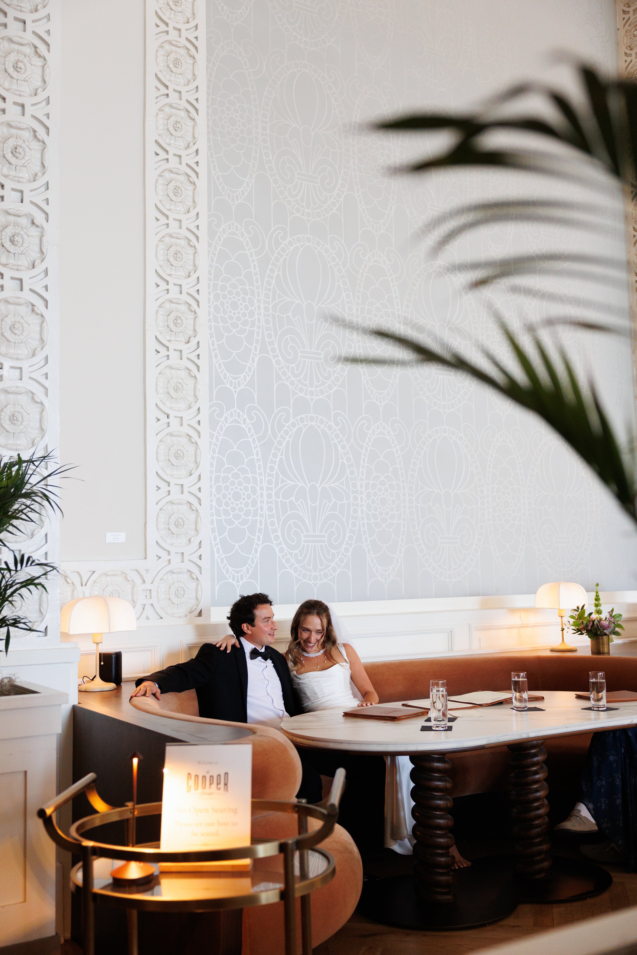 A couple in wedding attire sitting at a semi-circular booth in a decorated, elegant restaurant or event space. The man is wearing a tuxedo, and the woman is wearing a white gown, both smiling and appearing happy.