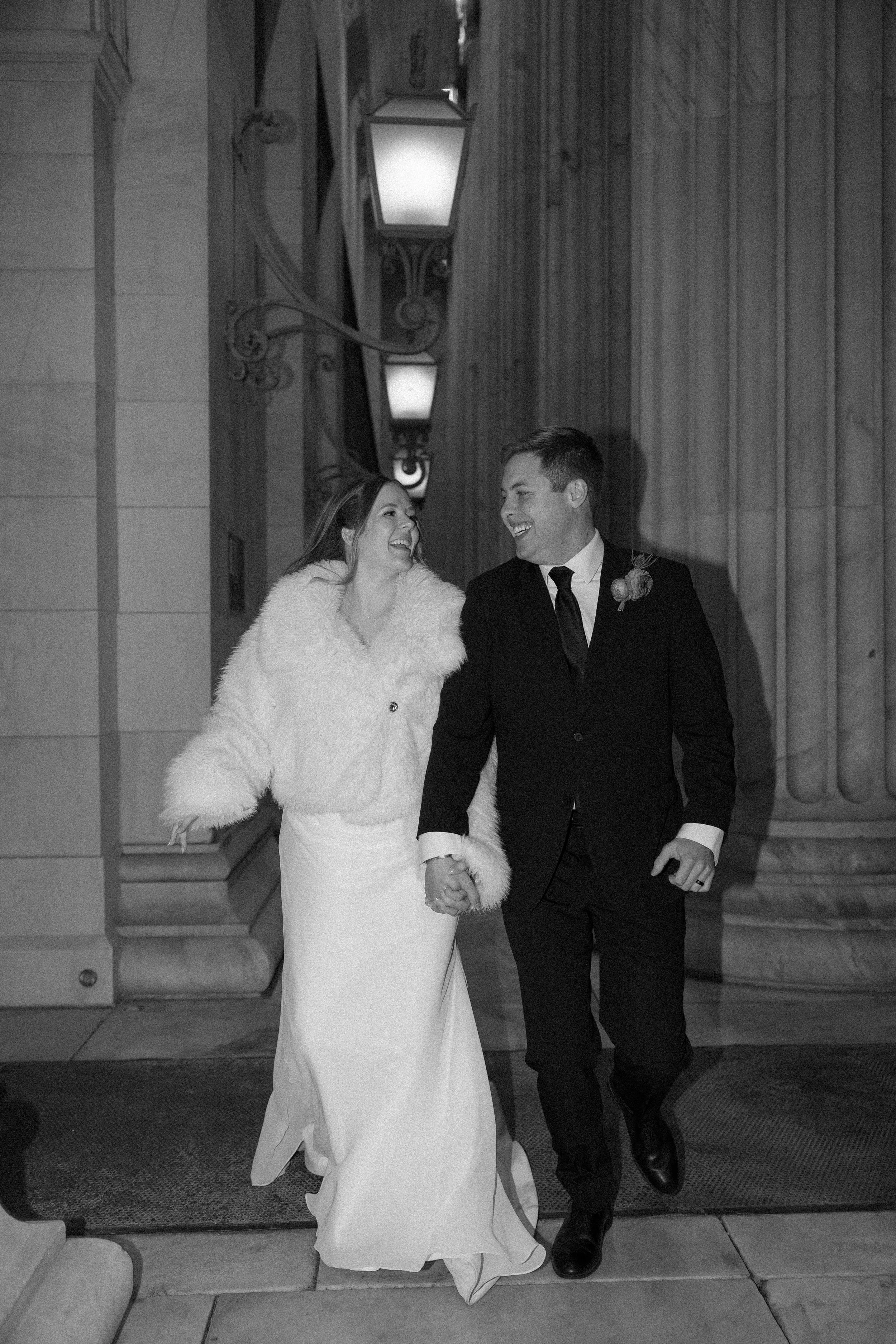 A smiling couple holding hands, walking outside at night, dressed in wedding attire, with the woman in a long white dress and furry white coat, and the man in a black suit and tie.