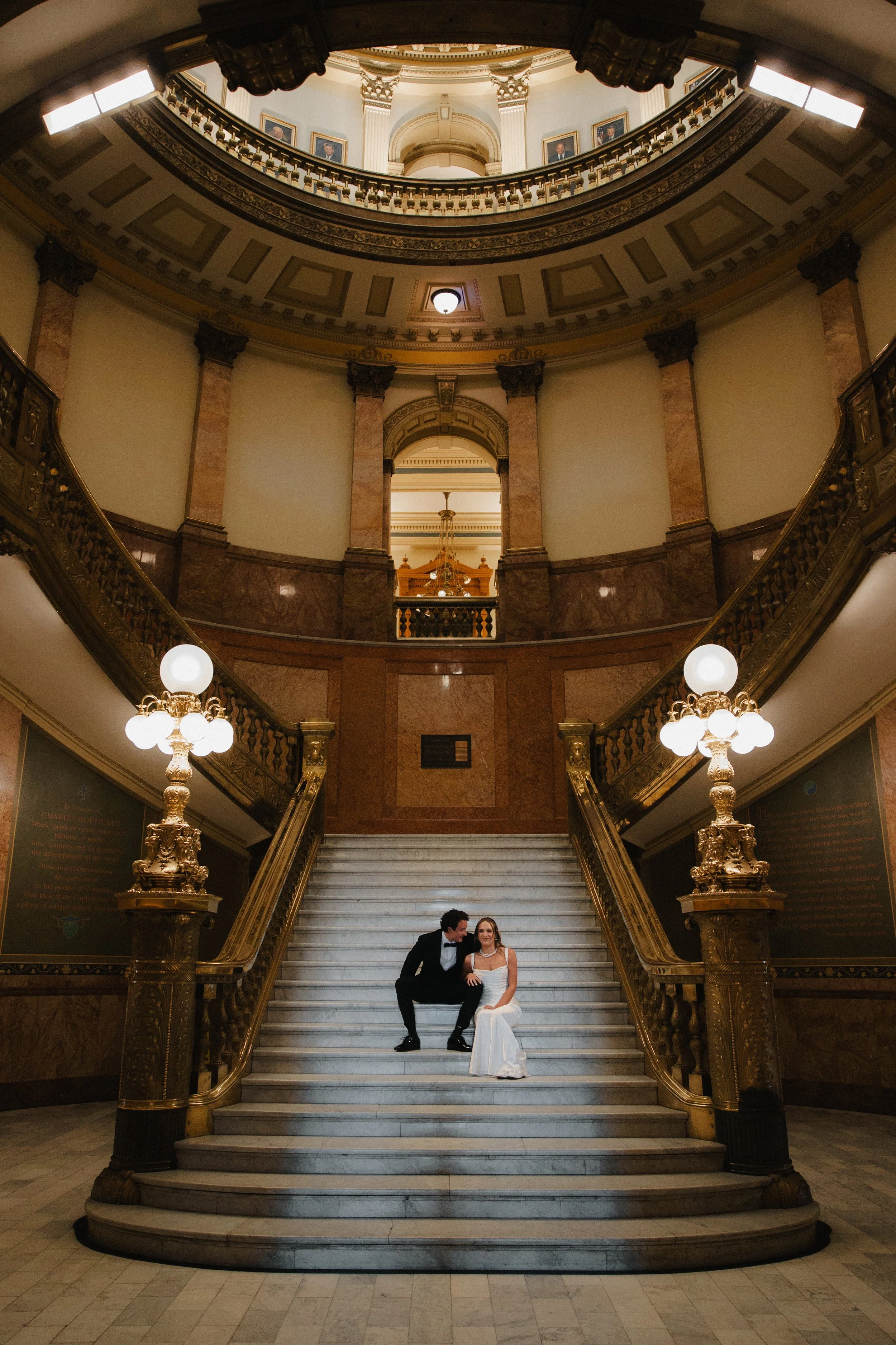 A couple dressed in formal attire sitting on a grand staircase inside an ornate, historic building with marble railings, decorative gold lamps, and a domed ceiling with portraits.