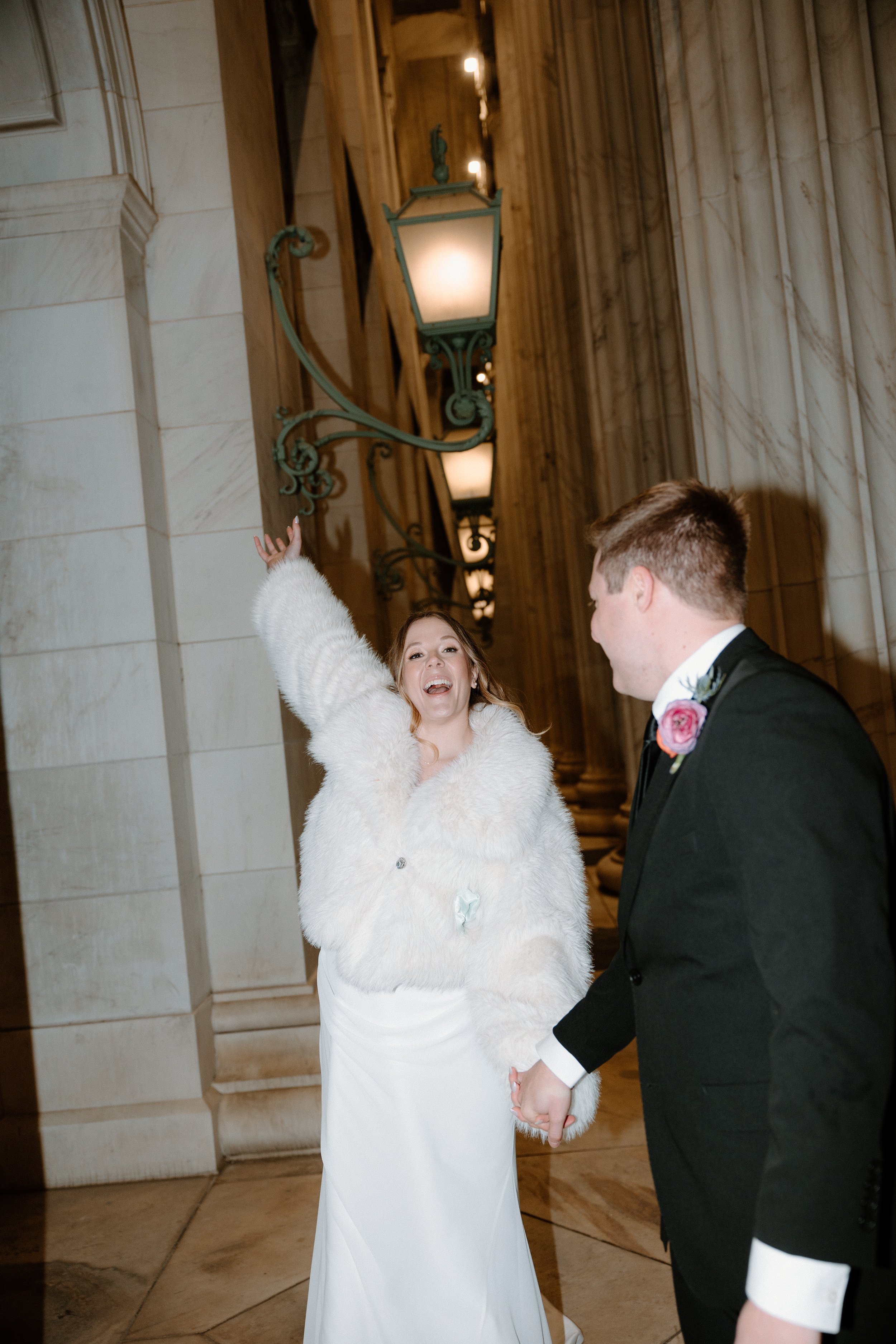 A joyous bride and groom holding hands and smiling at each other inside a grand, historic building with ornate lighting fixtures and marble walls.