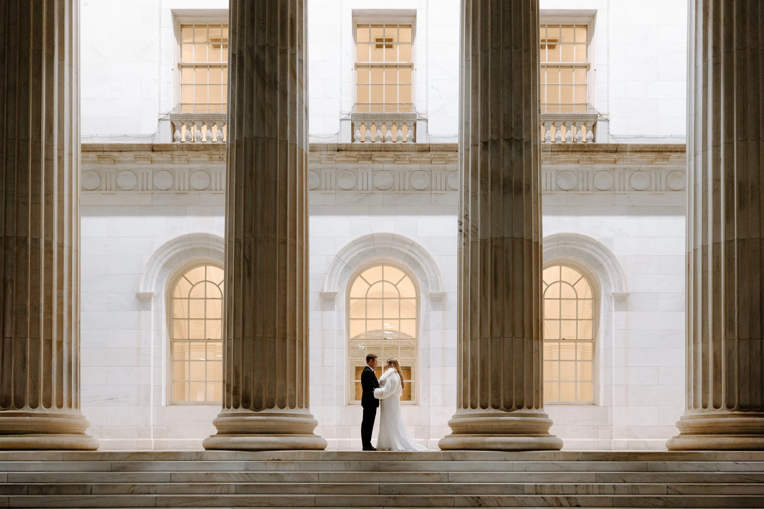 A bride and groom standing face-to-face in front of large classical columns, holding hands, inside a building with marble walls and arched windows.