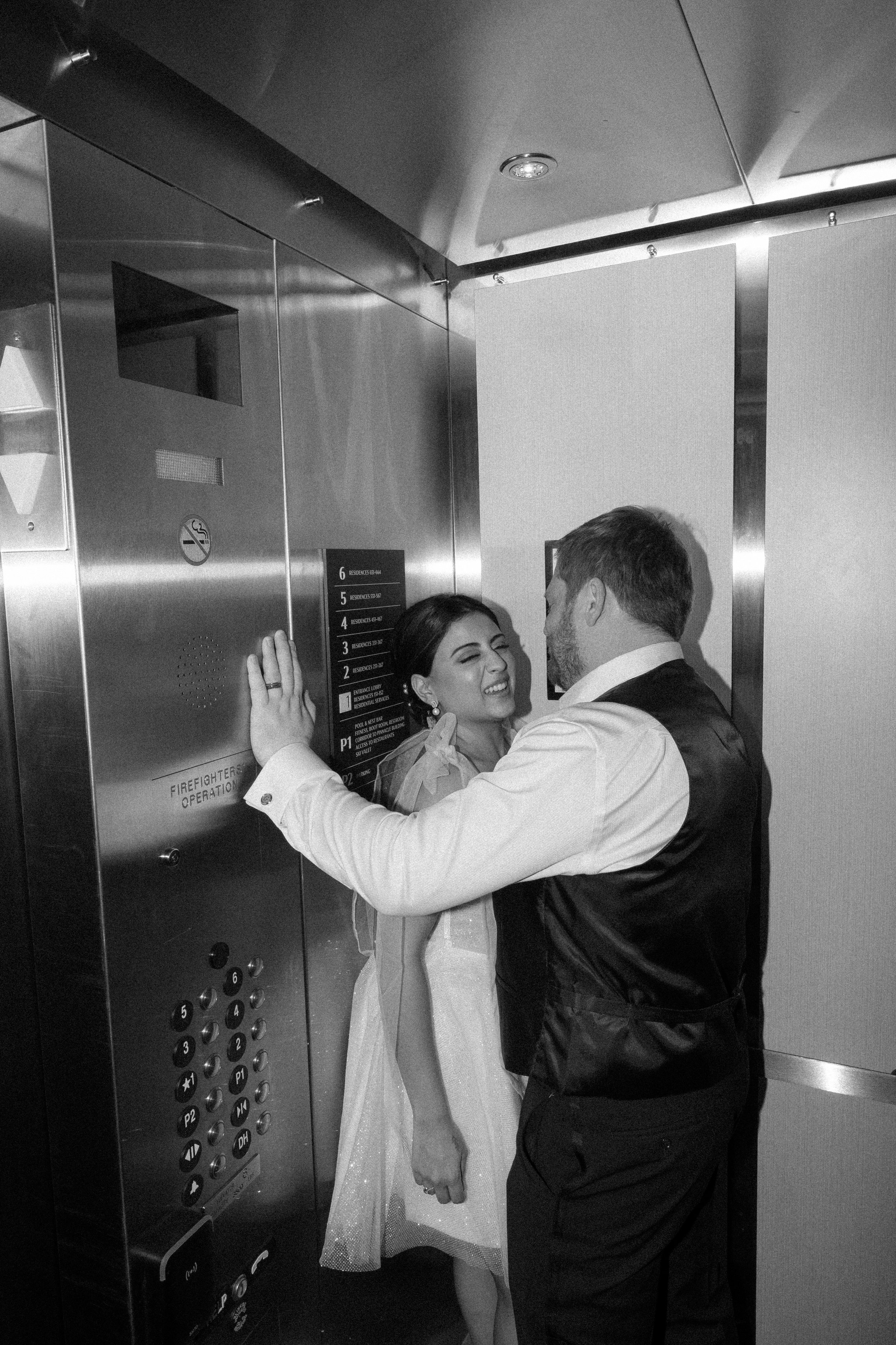 A man and woman sharing a moment inside an elevator. The woman is smiling, and the man is looking at her. They are close, with the man's hand on the elevator control panel. The woman is wearing a light-colored dress, and the man is dressed in a shirt