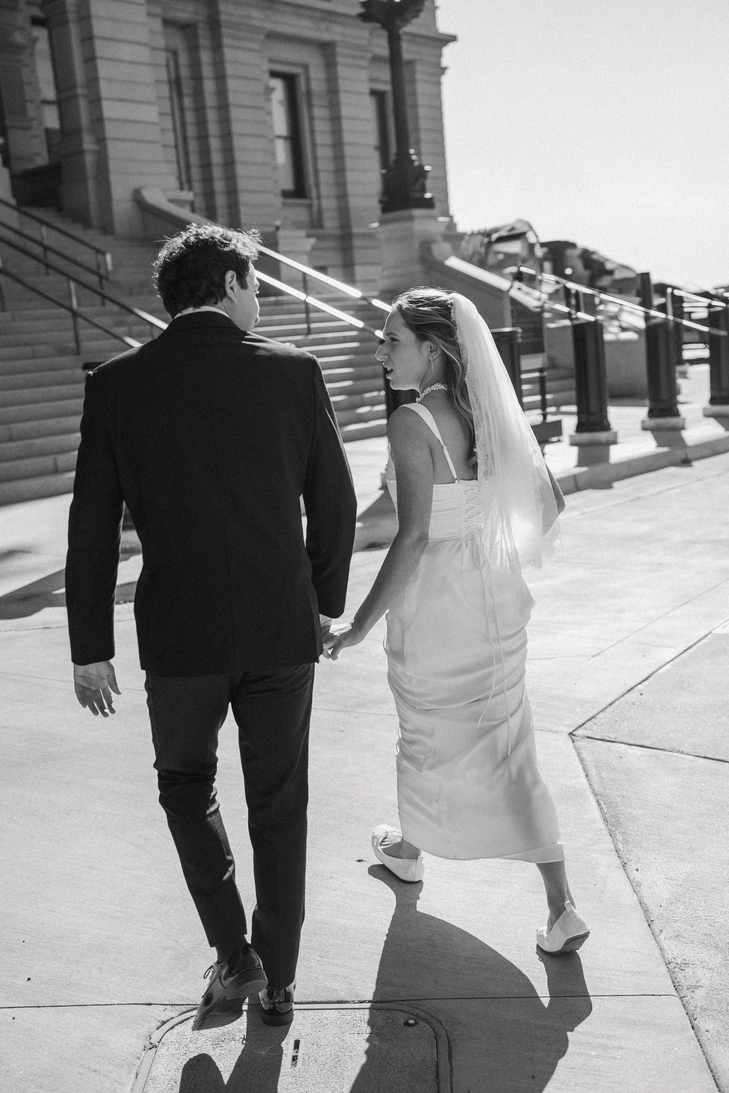 A black and white photo of a newlywed couple holding hands, walking outside in front of a building with stairs, on a sunny day.
