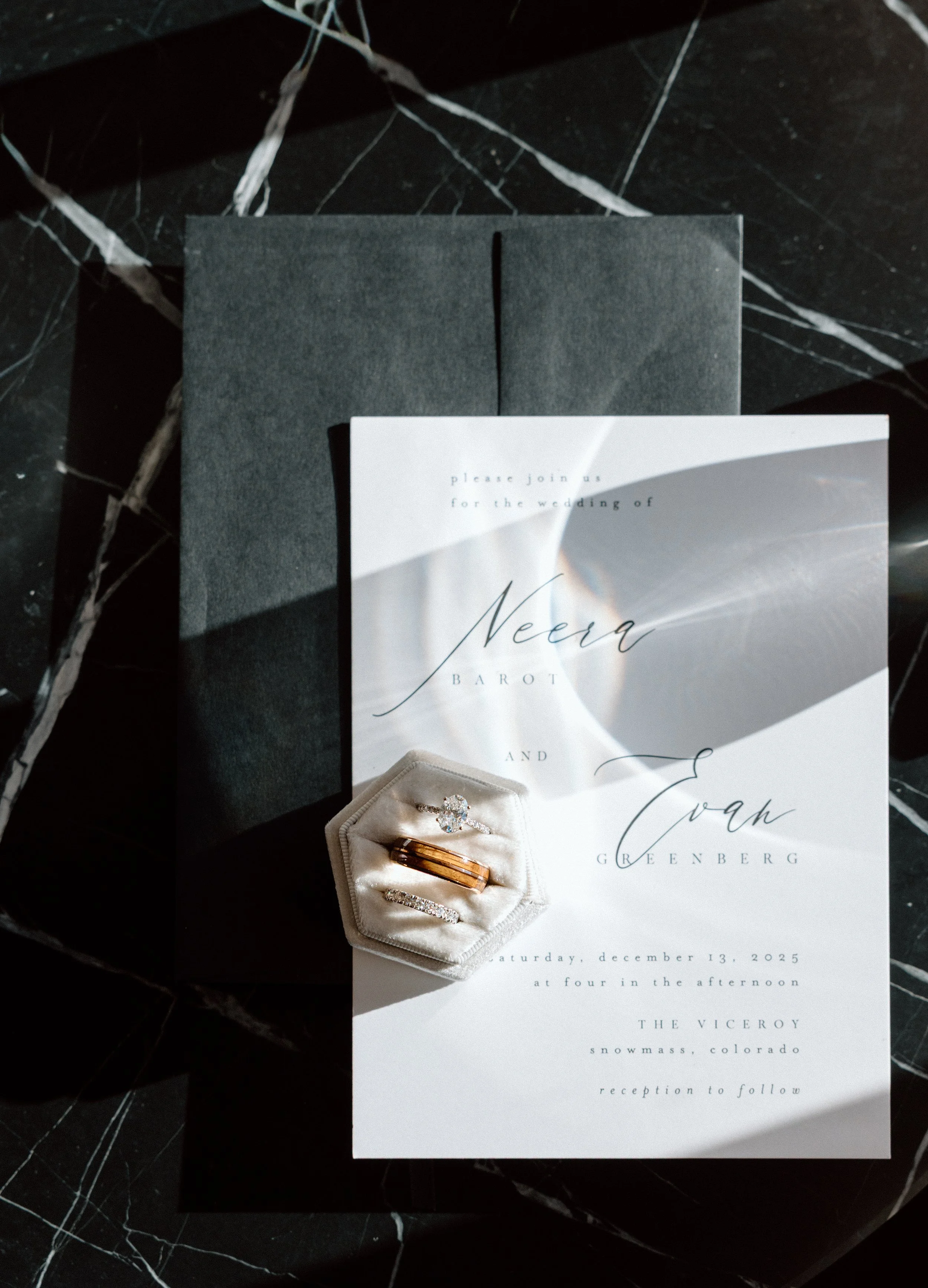 Wedding invitation with rings and an engagement ring on a black marble surface with white veining.