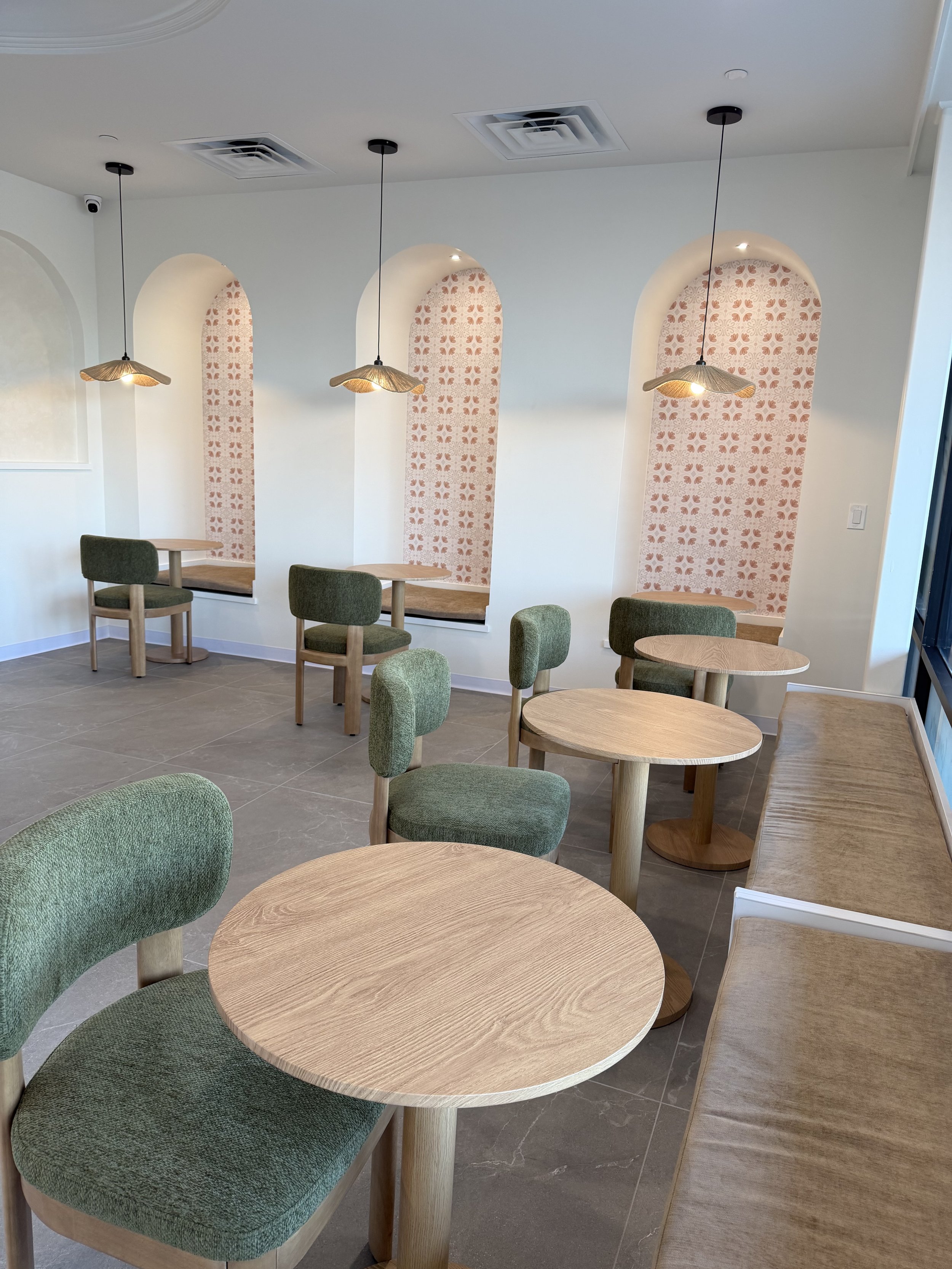 Interior of a modern cafe or restaurant with round wooden tables and green upholstered chairs. Decorated wall niches with patterned wallpaper and hanging pendant lights.