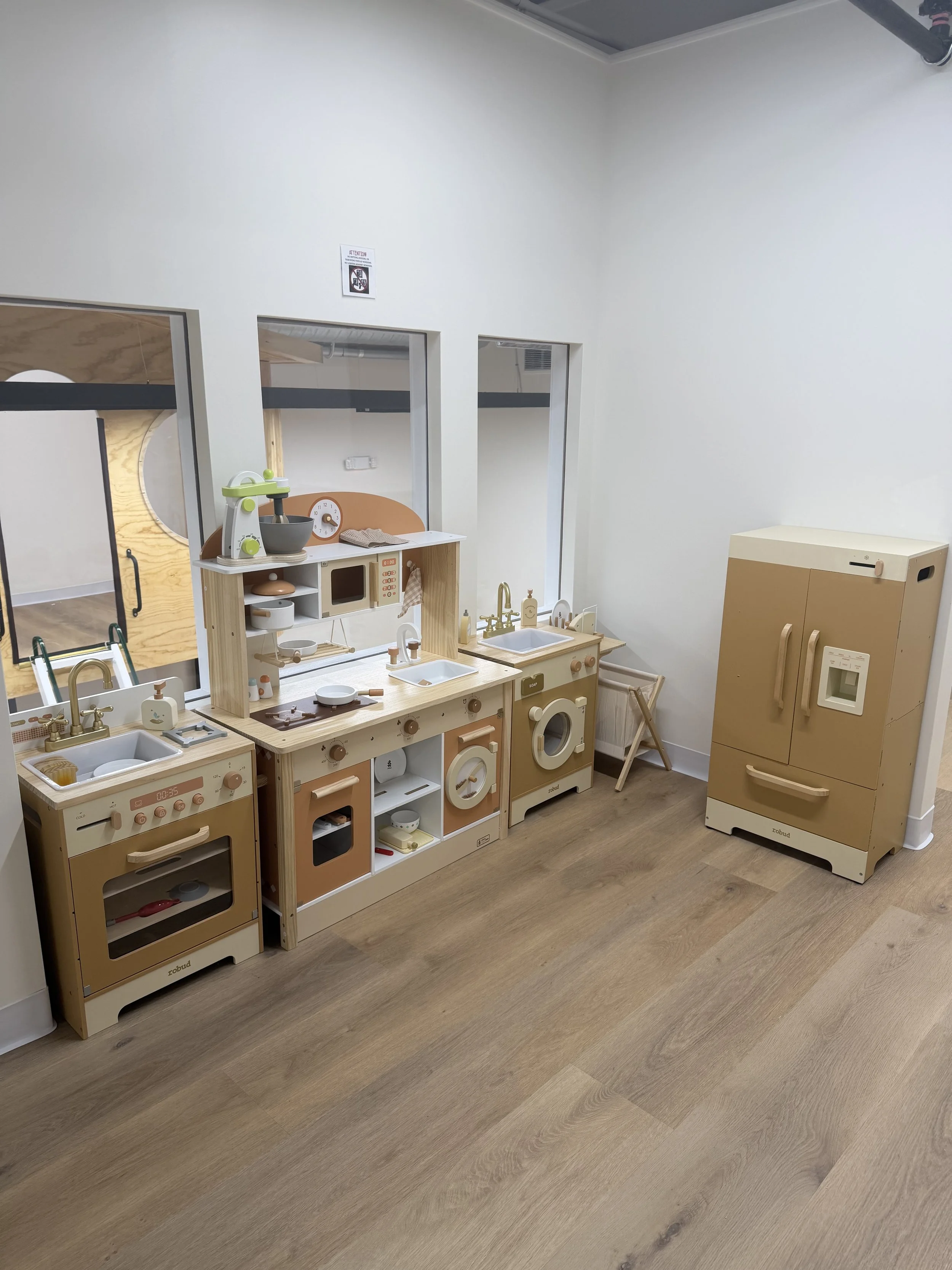Children's pretend play kitchen set with stove, microwave, sink, and storage cabinets, made of light wood and pastel accents, in a classroom setting.