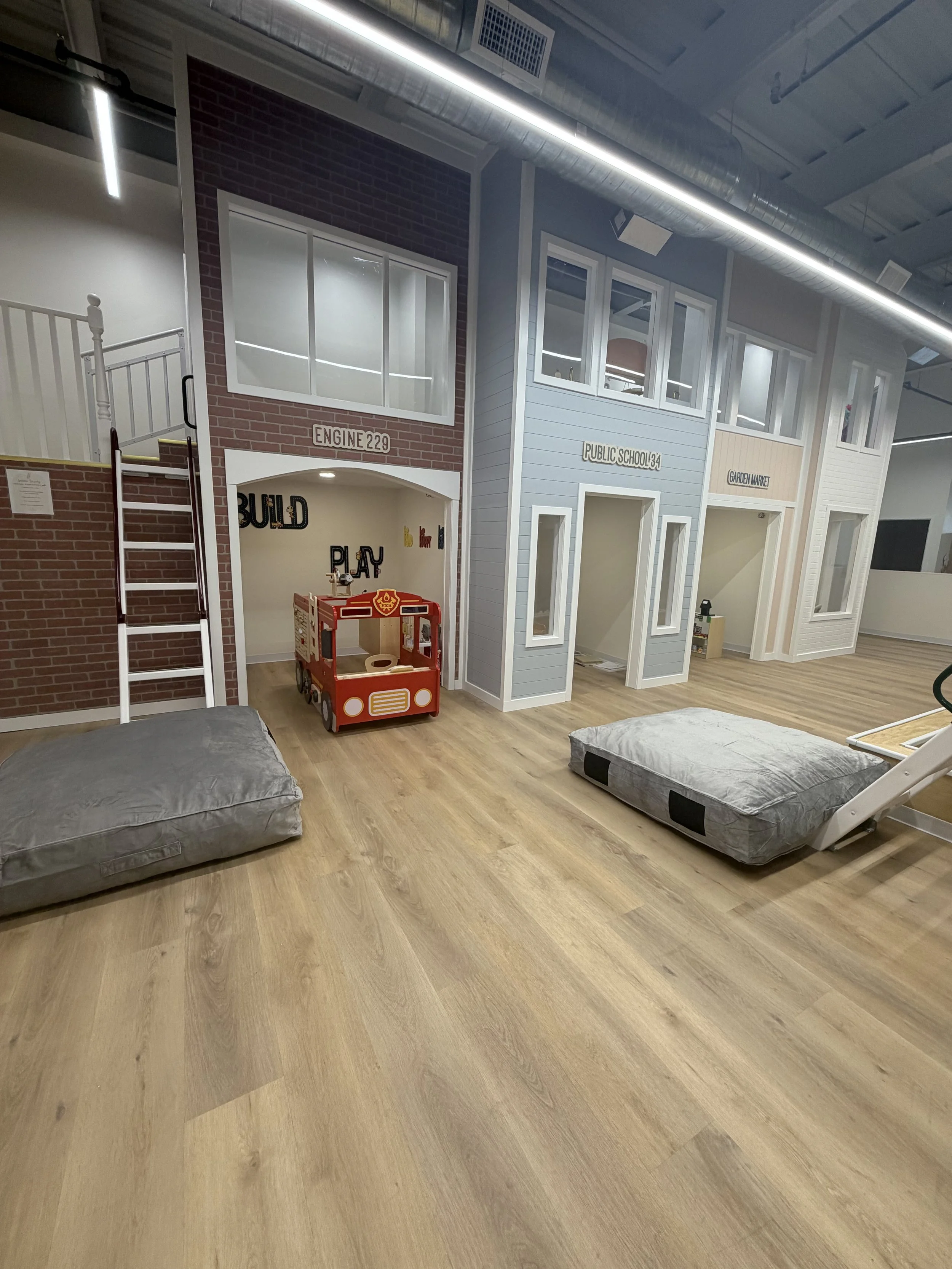Indoor play area designed to look like houses with signs indicating different rooms, a red toy bus, and foam mats on the floor.