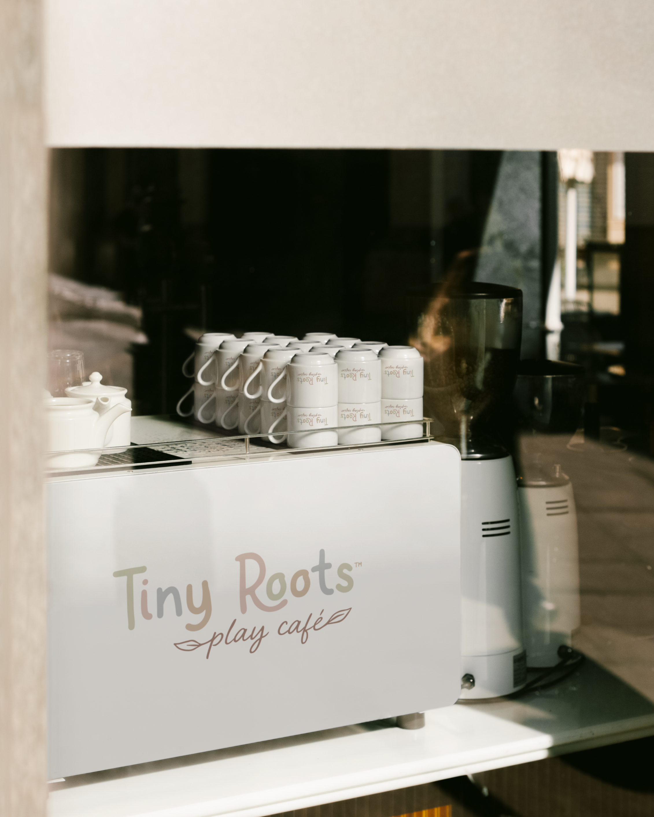 Inside a play café with a small coffee machine, several stacked white coffee mugs, a teapot, and a blender. The café sign reads 'Tiny Roots play café'.
