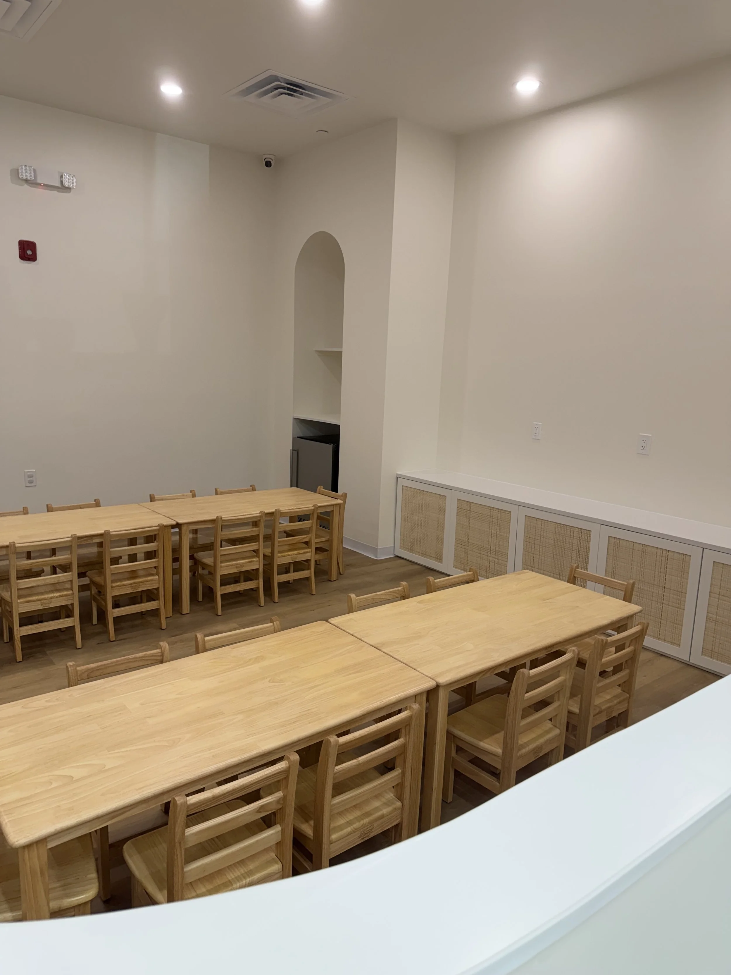 Empty room with light wood tables and chairs, white walls, recessed lighting, and built-in cabinets.