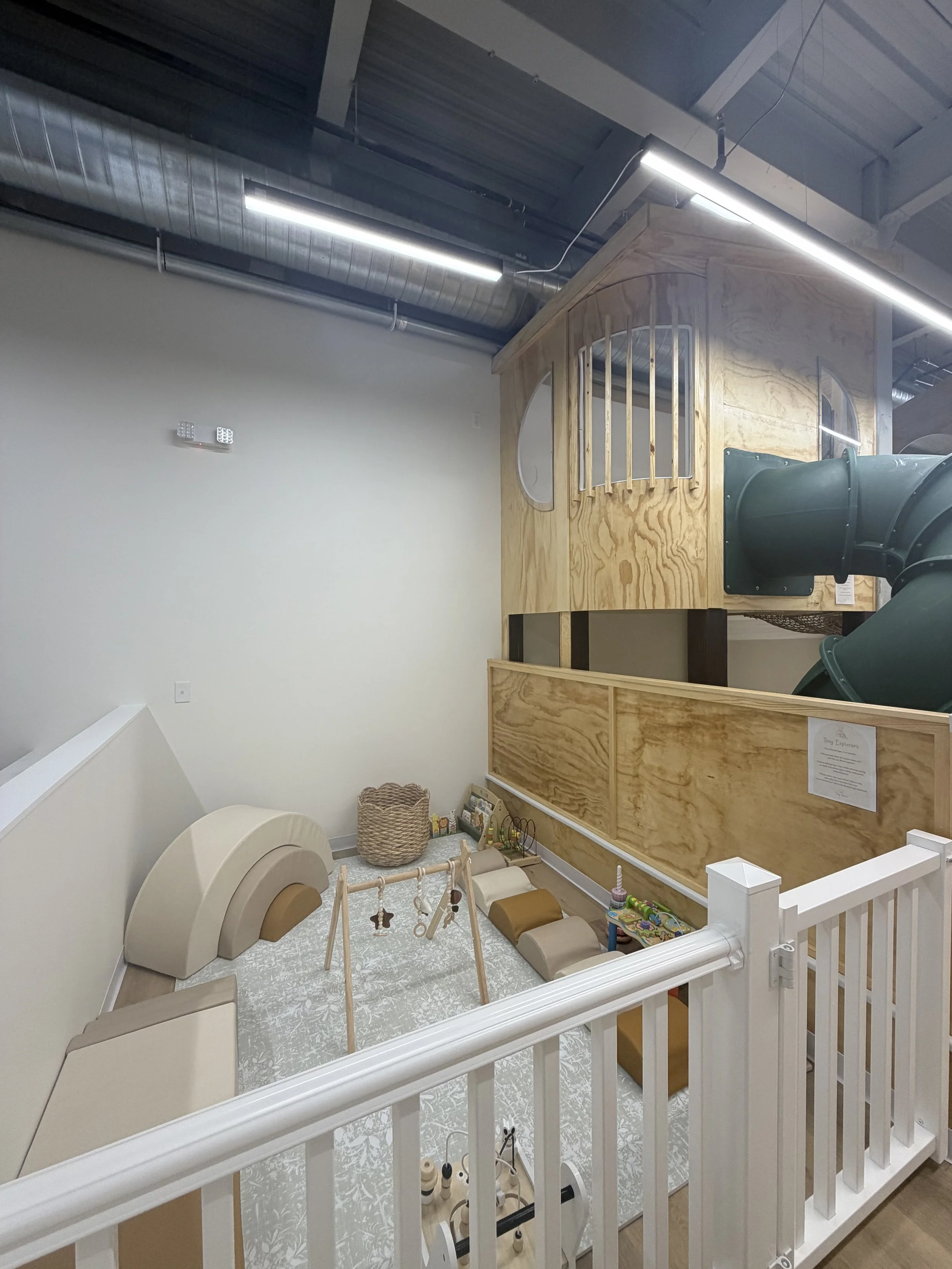 Indoor play area with wooden structure resembling a house, a green slide, and various soft toys and play equipment on a patterned rug, surrounded by white safety railing.