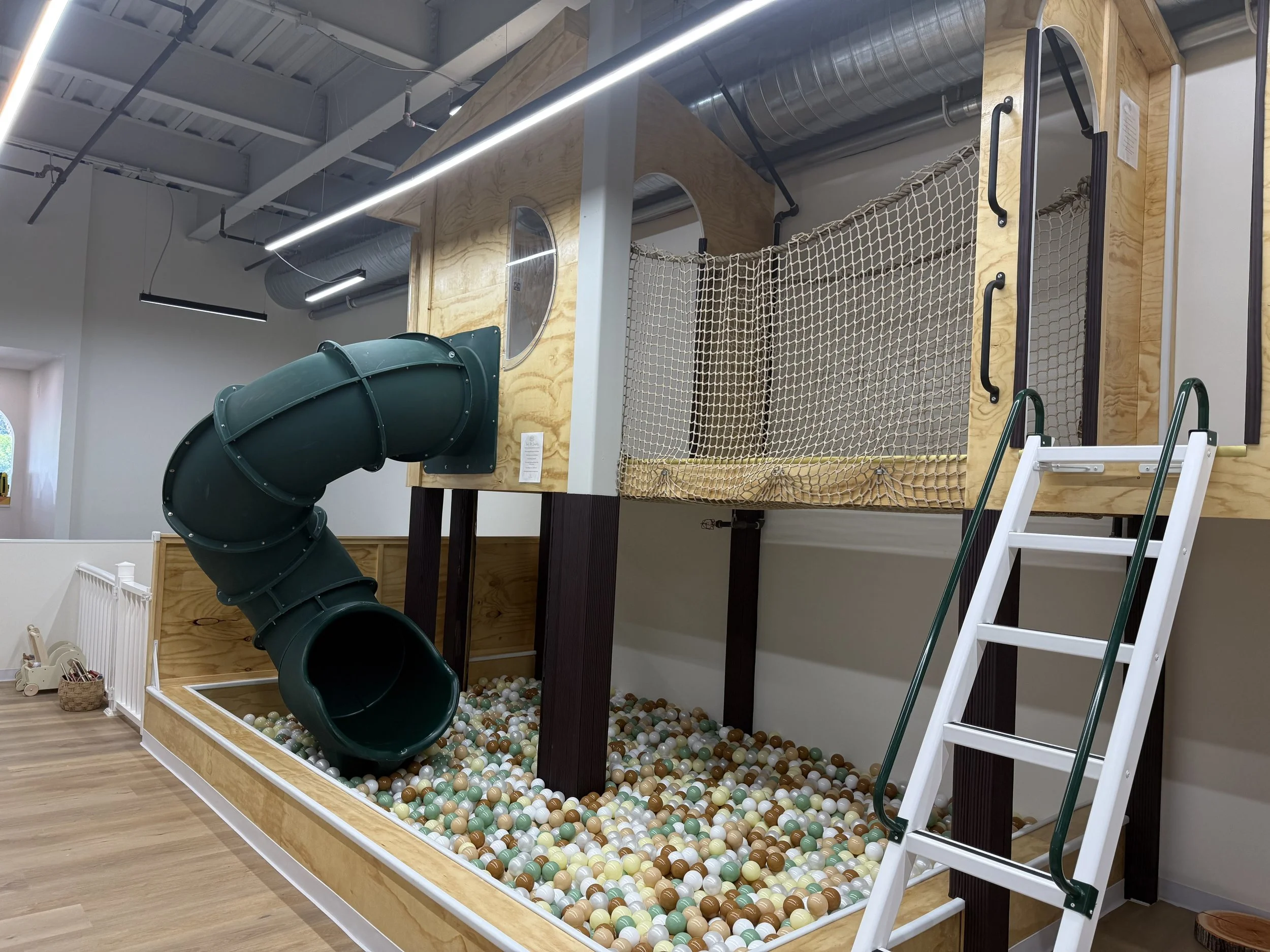An indoor playground with a green spiral slide leading into a ball pit filled with multi-colored plastic balls, surrounded by wooden framing and netting.