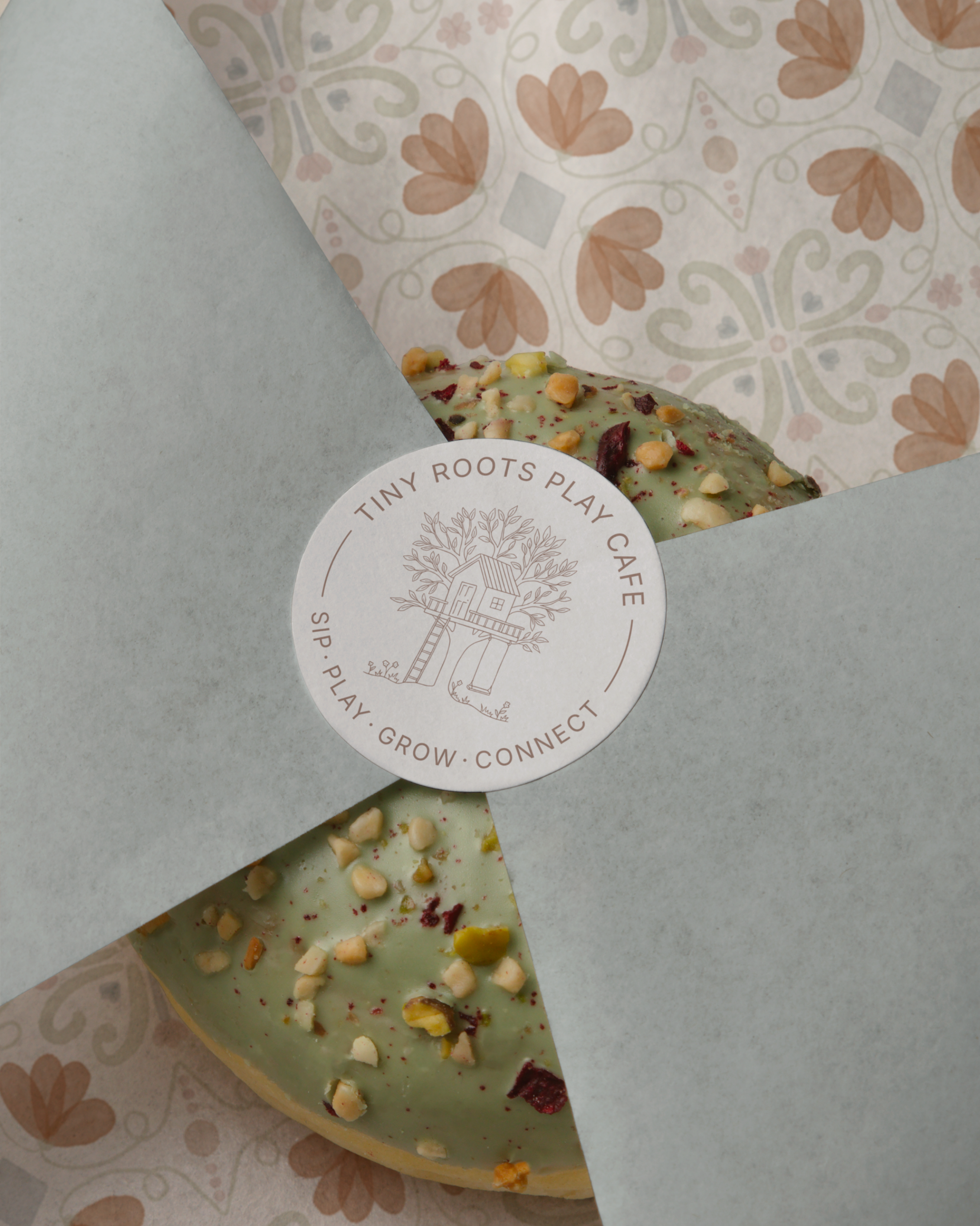 A partially unwrapped green tea flavored pastry with chopped nuts and dried berries on top, in paper wrapping with a circular logo sticker from Tiny Roots Play Cafe.