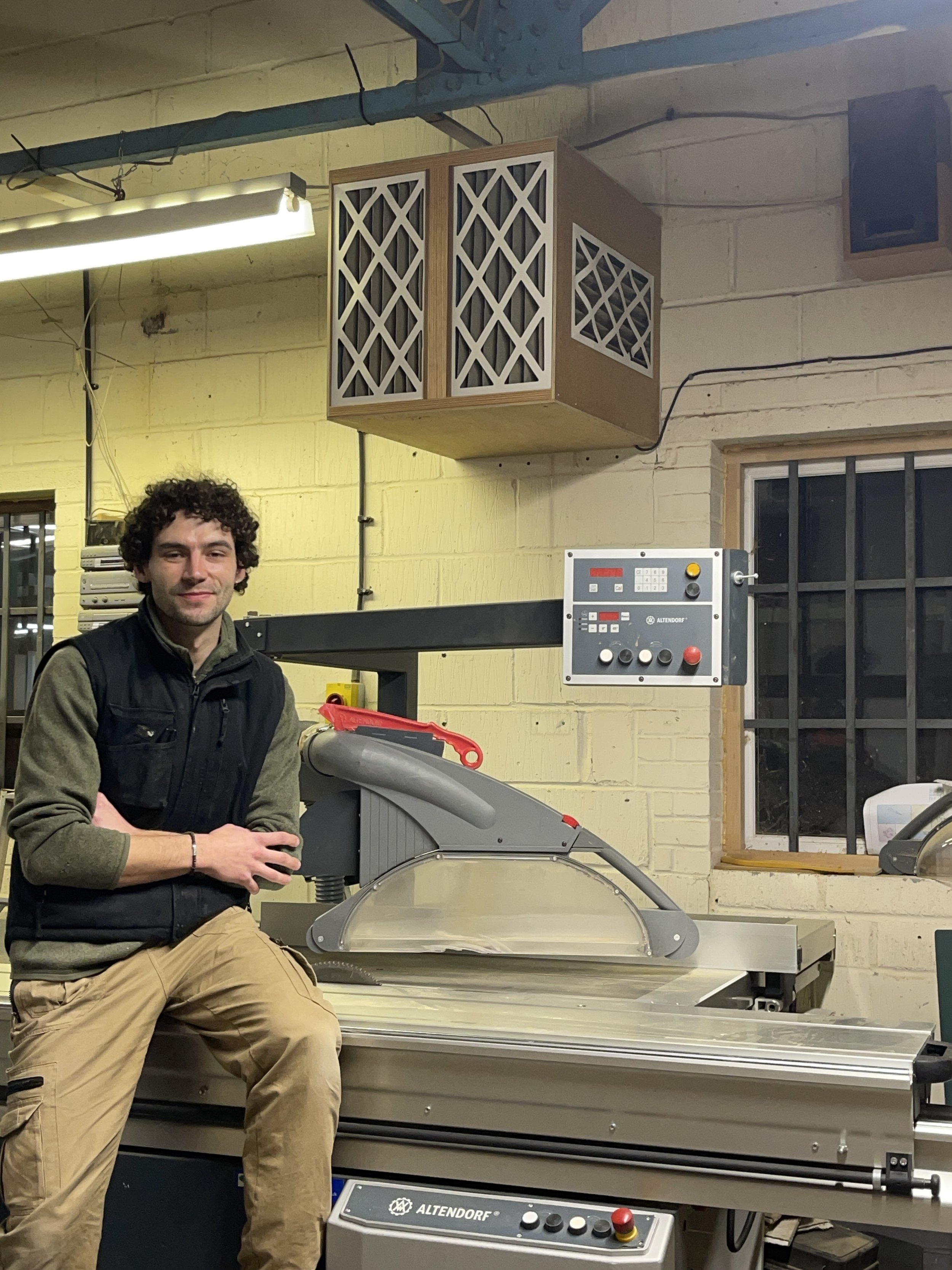 A young man with curly hair wearing a black vest and khaki pants standing next to a large industrial saw in a workshop with yellow brick walls, a window, and a control panel.