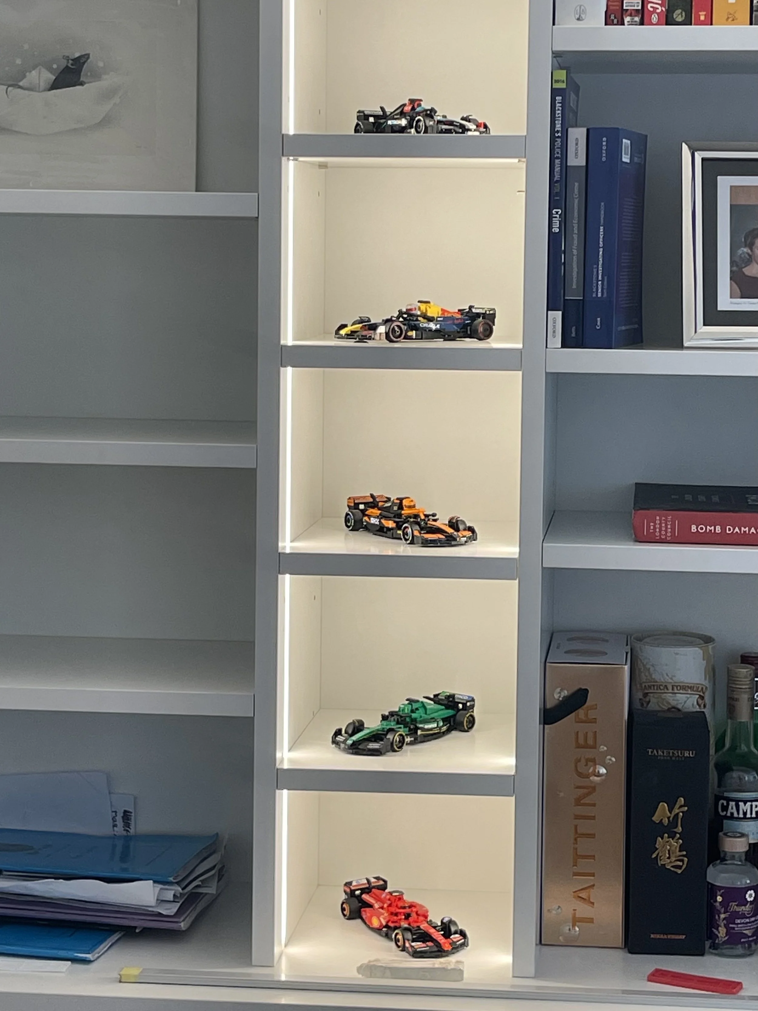 Shelf with five model Formula 1 cars lined up vertically, displayed in illuminated cubicles, with books and other items surrounding the shelf.