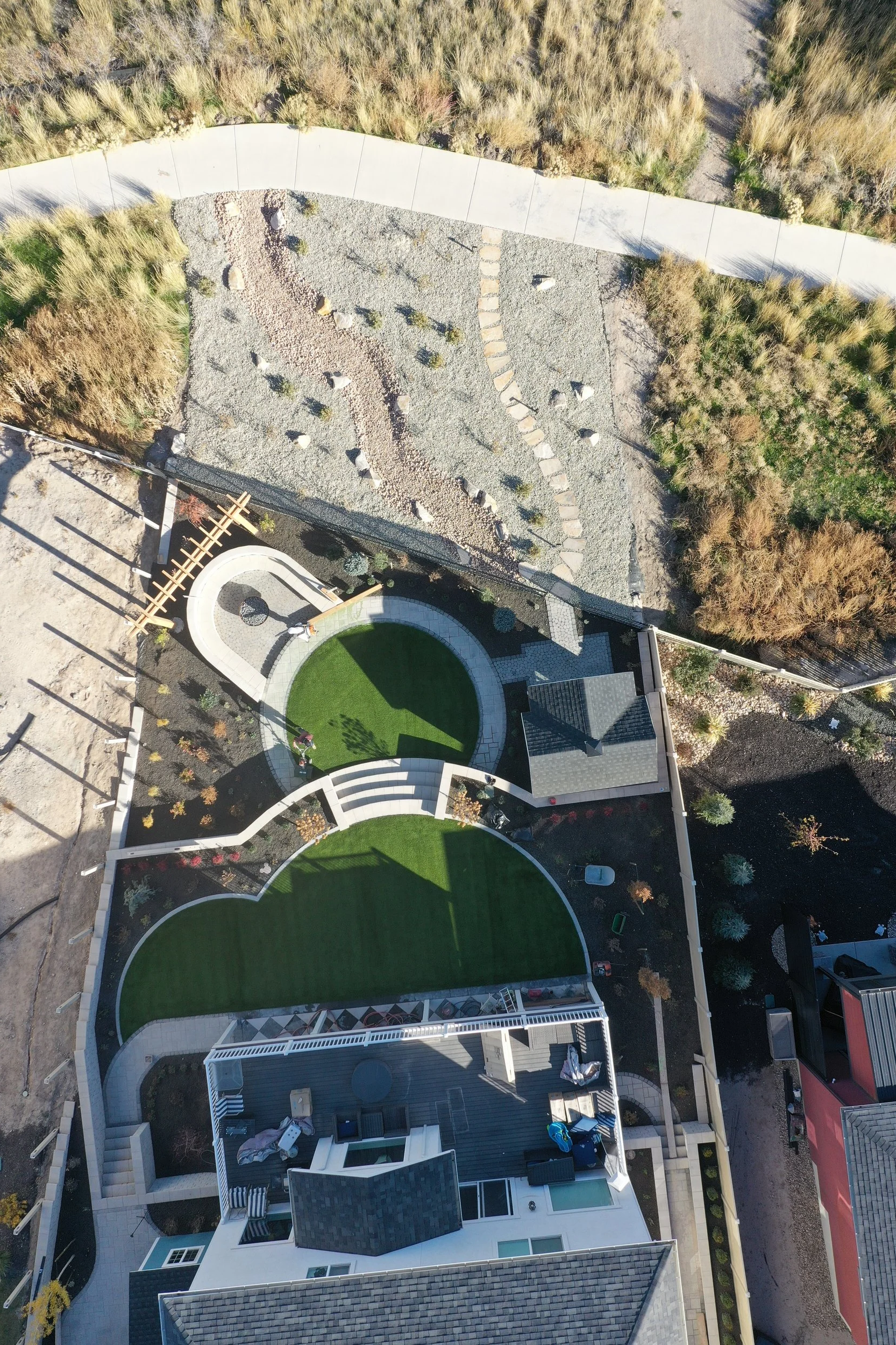 An aerial view of a backyard with a manicured lawn, concrete pathways, small plants, and a deck, bordered by natural grassy areas and trees.