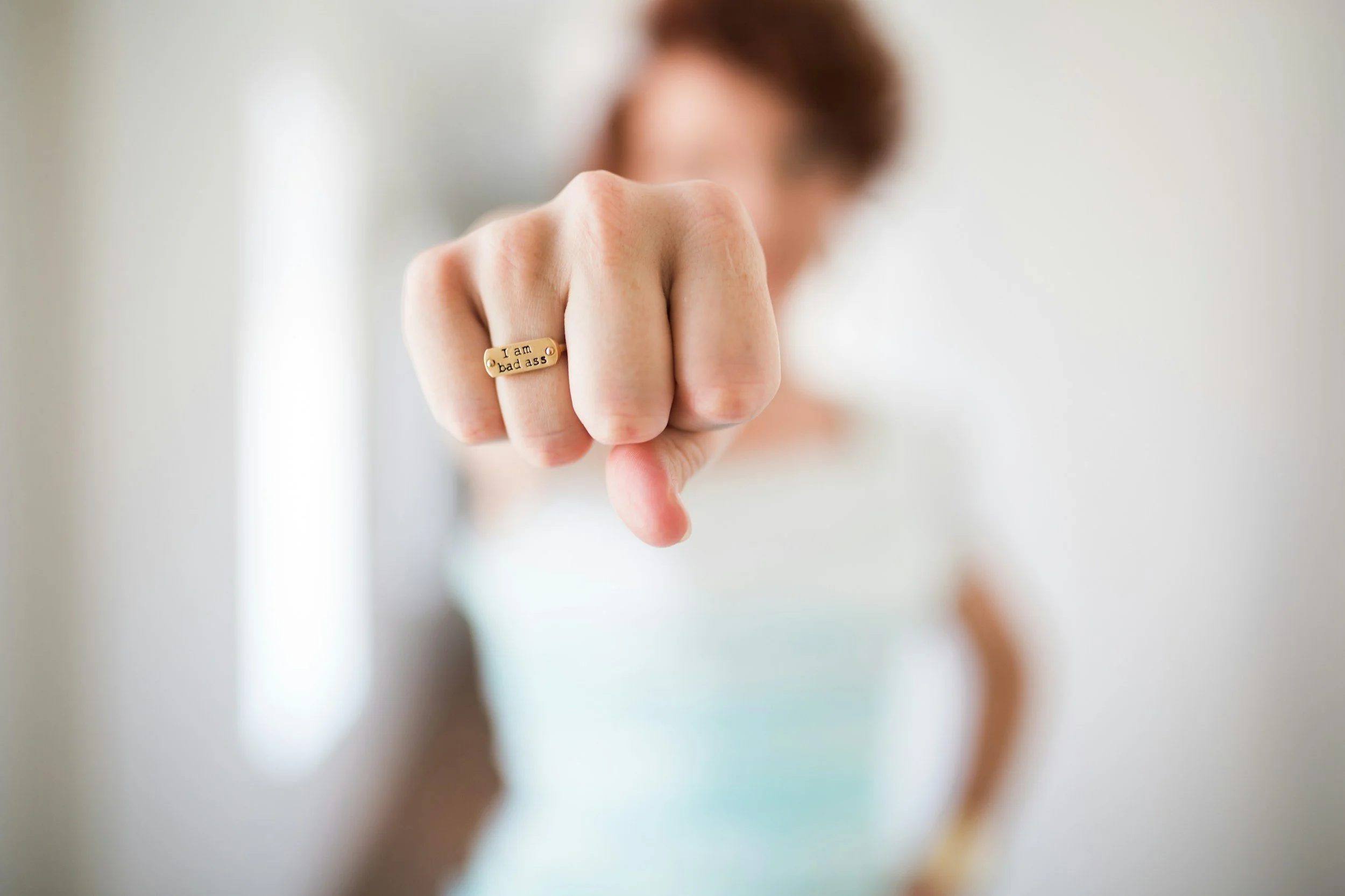 Close-up of a woman's hand with a gold ring that reads 'I am bad ass', pointing directly at the camera, blurred background.