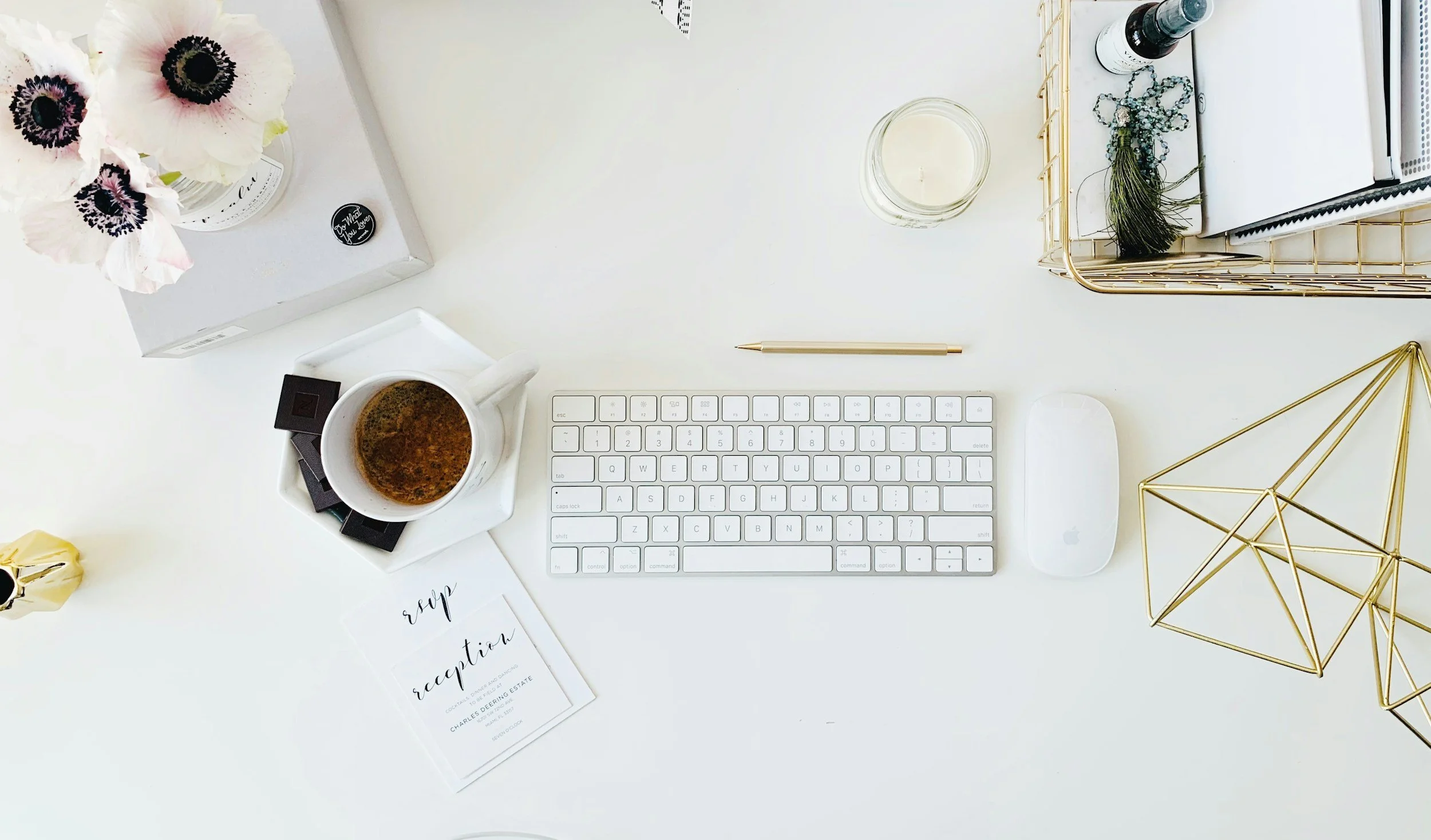 Top-down view of a clean white desk with a keyboard, mouse, pen, candle, cup of coffee, decorative gold geometric sculptures, a tray with office supplies, a small jar, a bouquet of pink and white flowers, and a receipt.