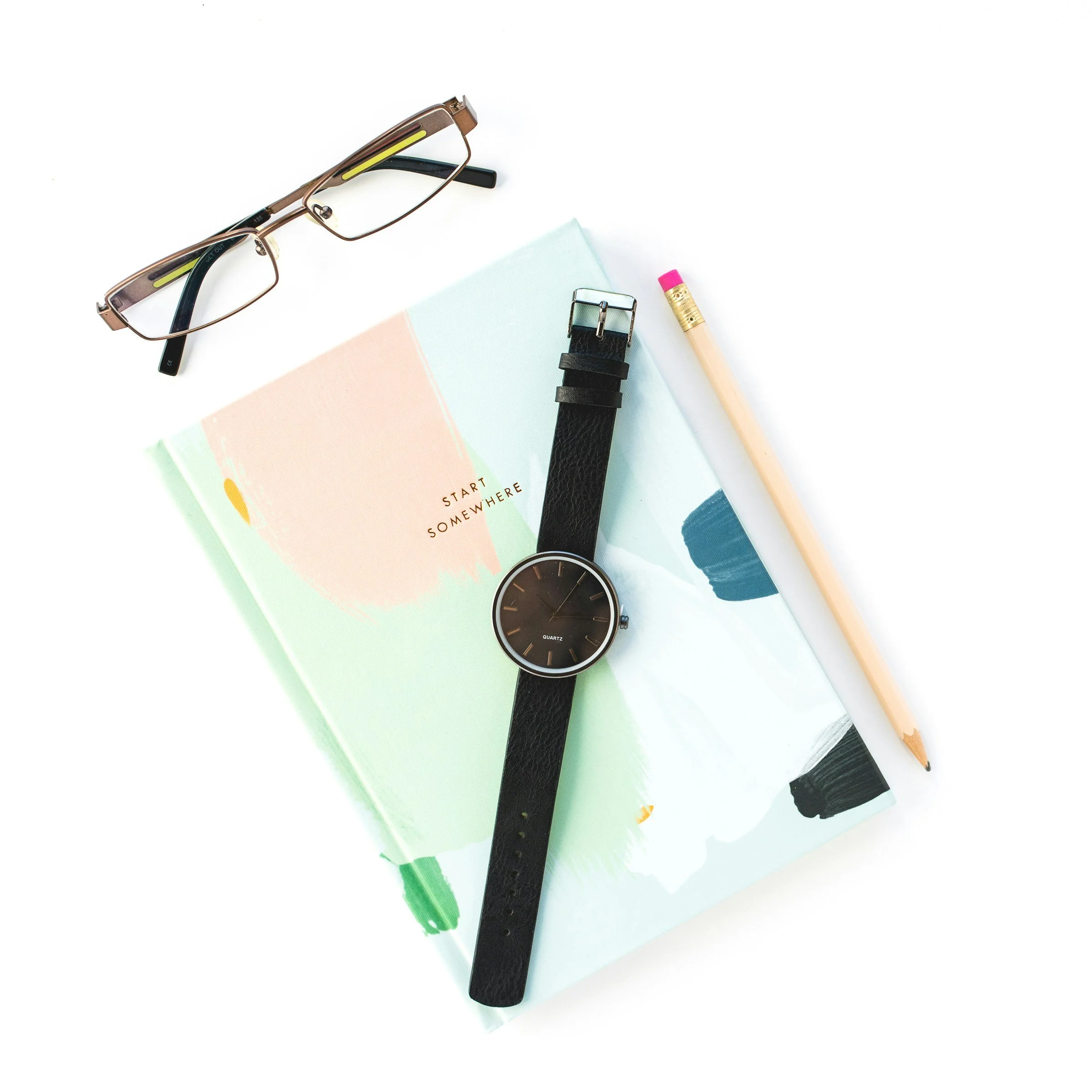 Eyeglasses, a black wristwatch, a pink eraser, a wooden pencil, and a pastel-colored book with abstract artwork on a white background.