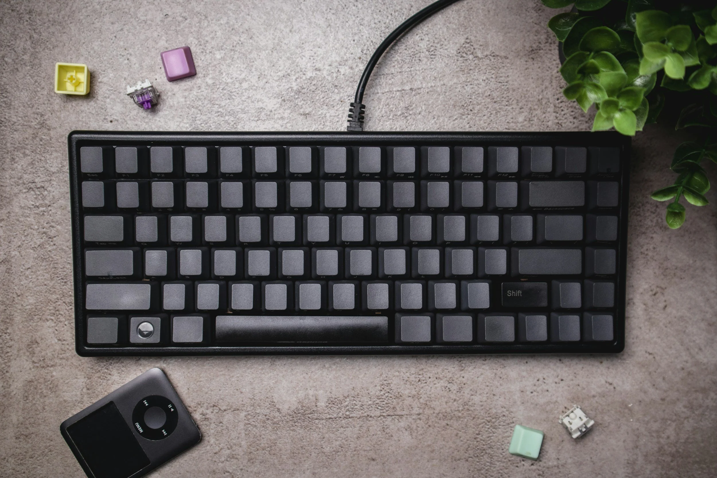 A black computer keyboard on a beige textured surface, with a small electronic device resembling an iPod Nano below it, and colorful keyboard switches and a green plant in the background.