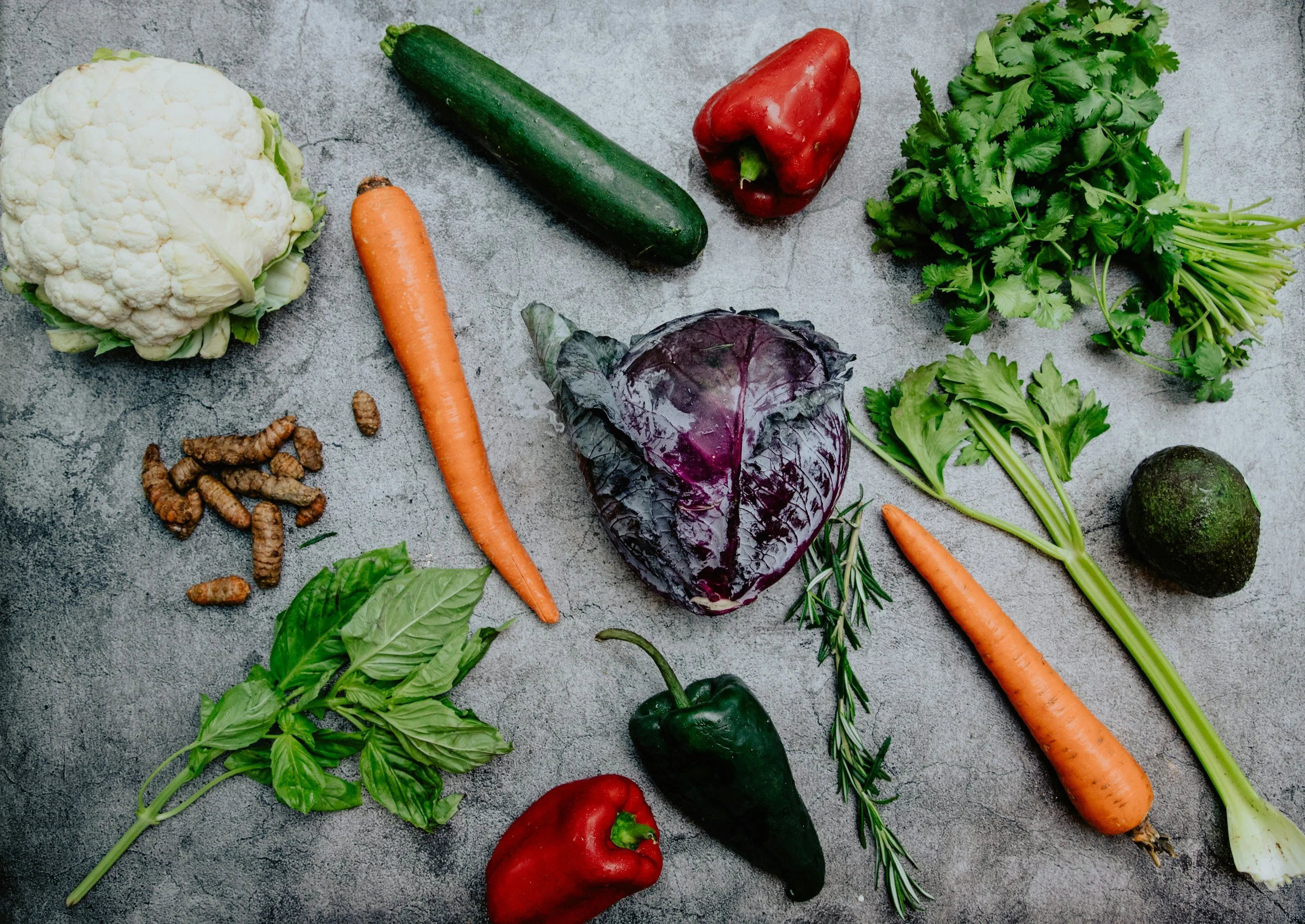 Assorted fresh vegetables including cauliflower, carrots, cucumbers, red and green bell peppers, cilantro, basil, an avocado, a purple cabbage, and sprigs of rosemary on a gray surface.