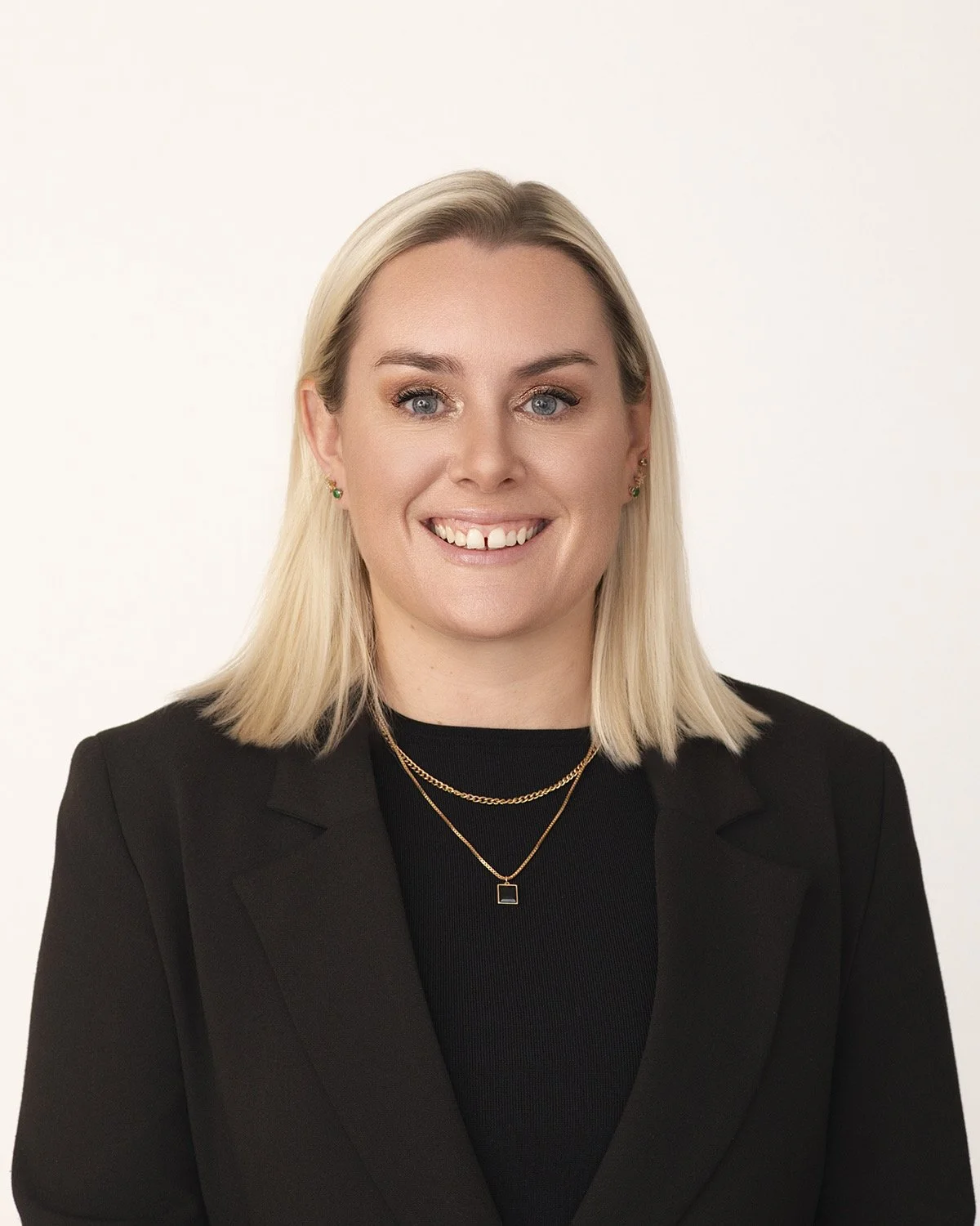 A woman with blonde hair, wearing a black blazer and black top, smiling against a plain white background.