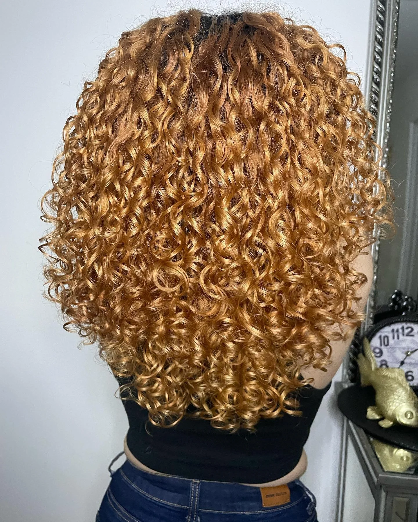 Back view of a woman with long layers ,3A curly, reddish-blonde hair, wearing a black top and blue jeans.