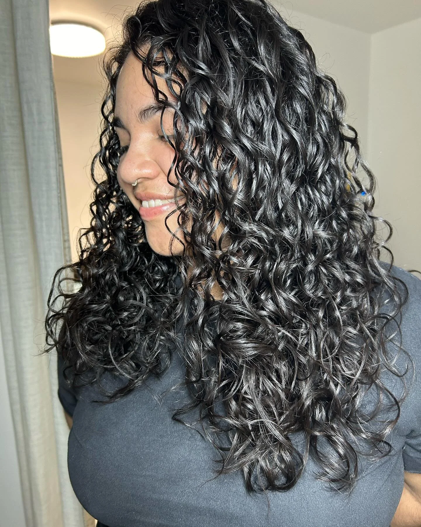 A woman with long, 3A curly black hair with curly face framing pieces, smiling with eyes closed, wearing a septum ring and a dark gray top, indoors.