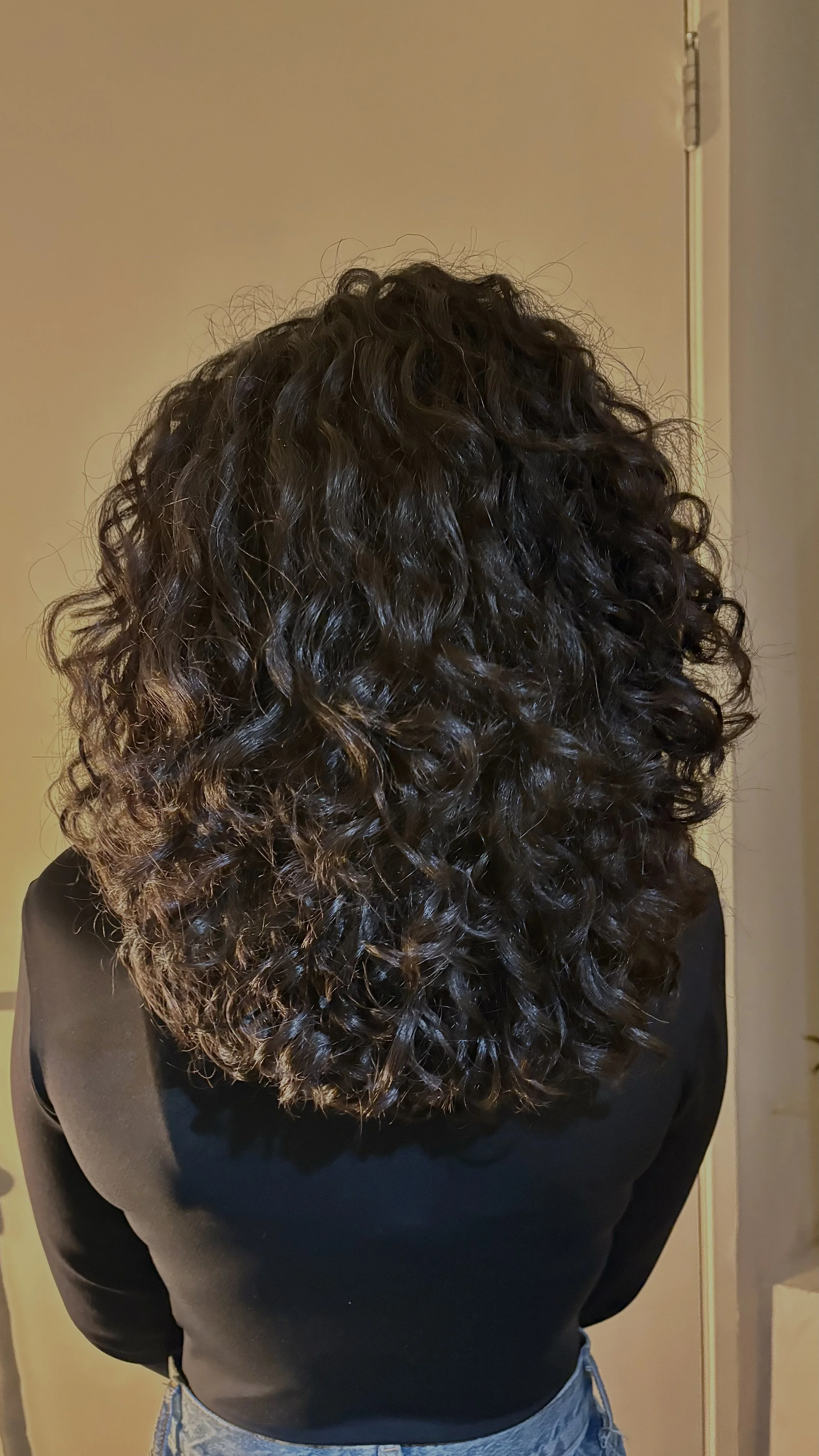 Back of a woman with voluminous, 3A curly dark hair, short round layers wearing a black top and blue jeans.
