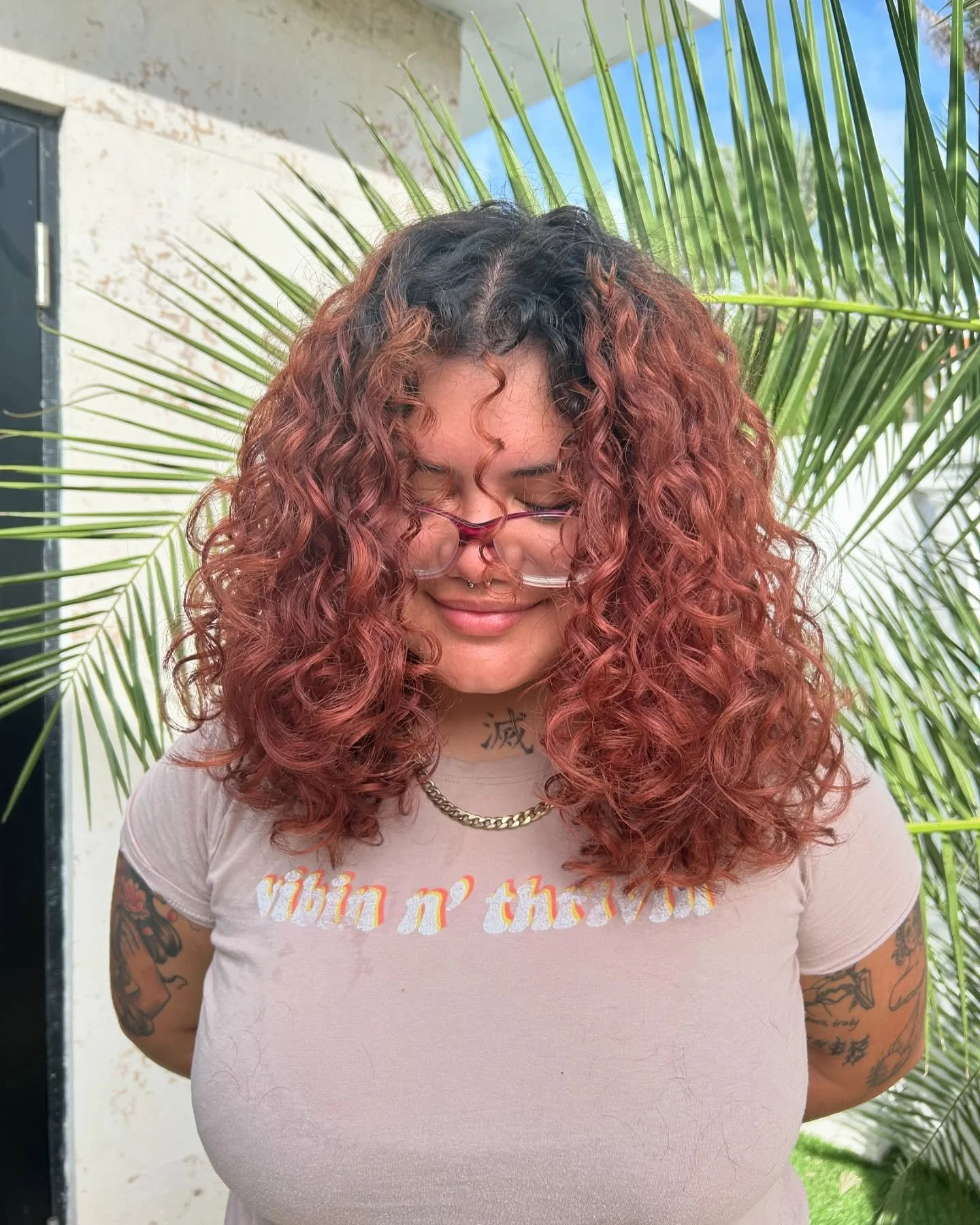 A woman with 3A curly, round layers and reddish-pink hair standing outdoors in front of a green palm tree, smiling with her eyes closed, wearing glasses, a necklace, and a light-colored T-shirt.