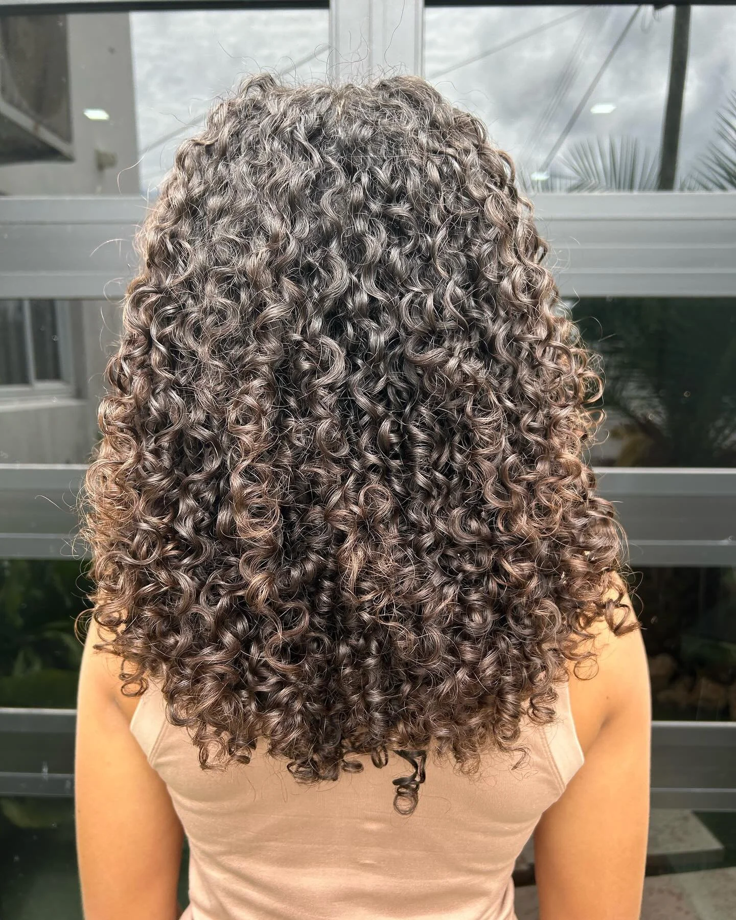 A woman with dark brown, tight, 3B 3C short layers curly hair standing outdoors in front of a glass window.