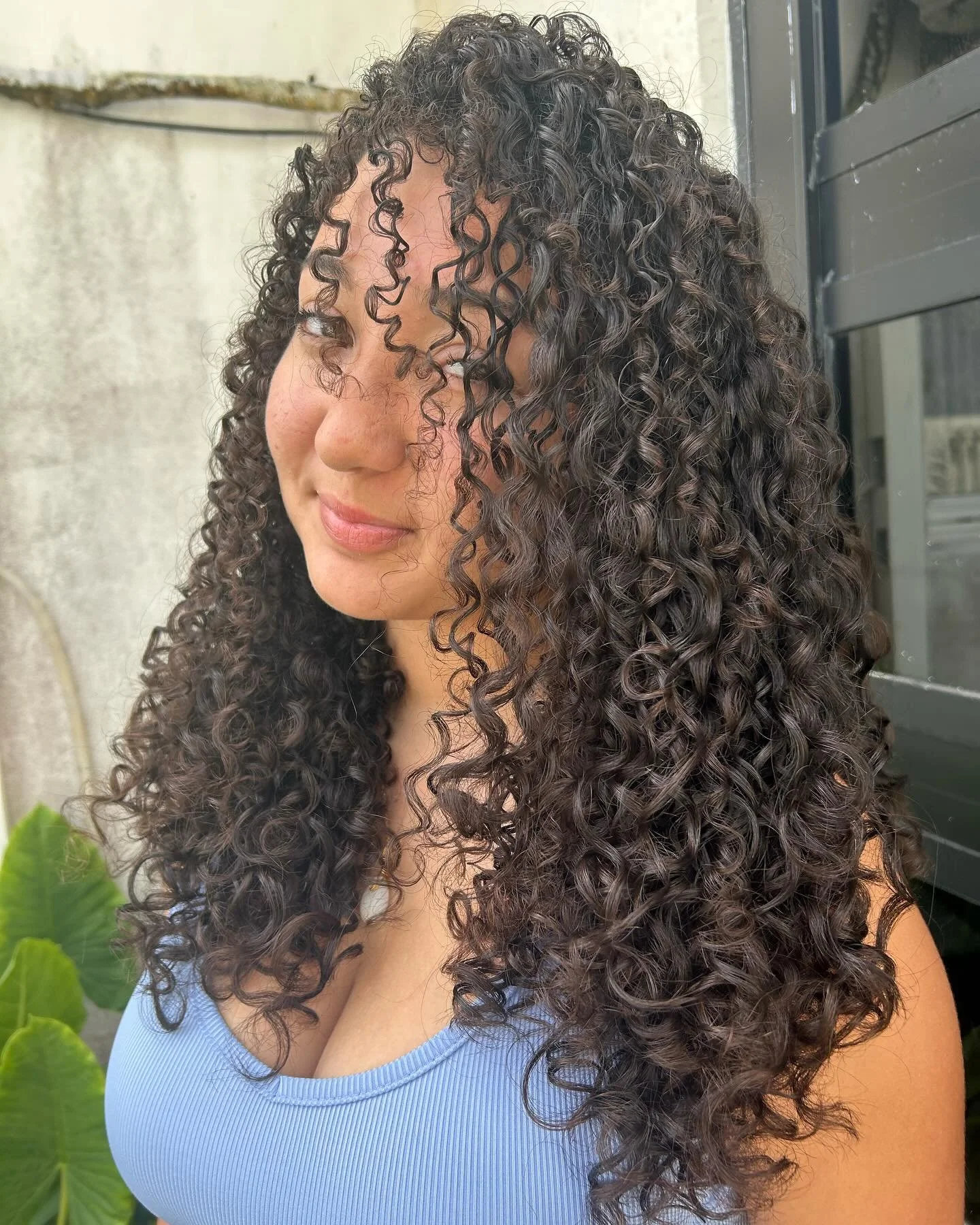 A woman with long, dark, 3B curly hair smiling softly, wearing a light blue top, standing outdoors near a wall and some green leaves.