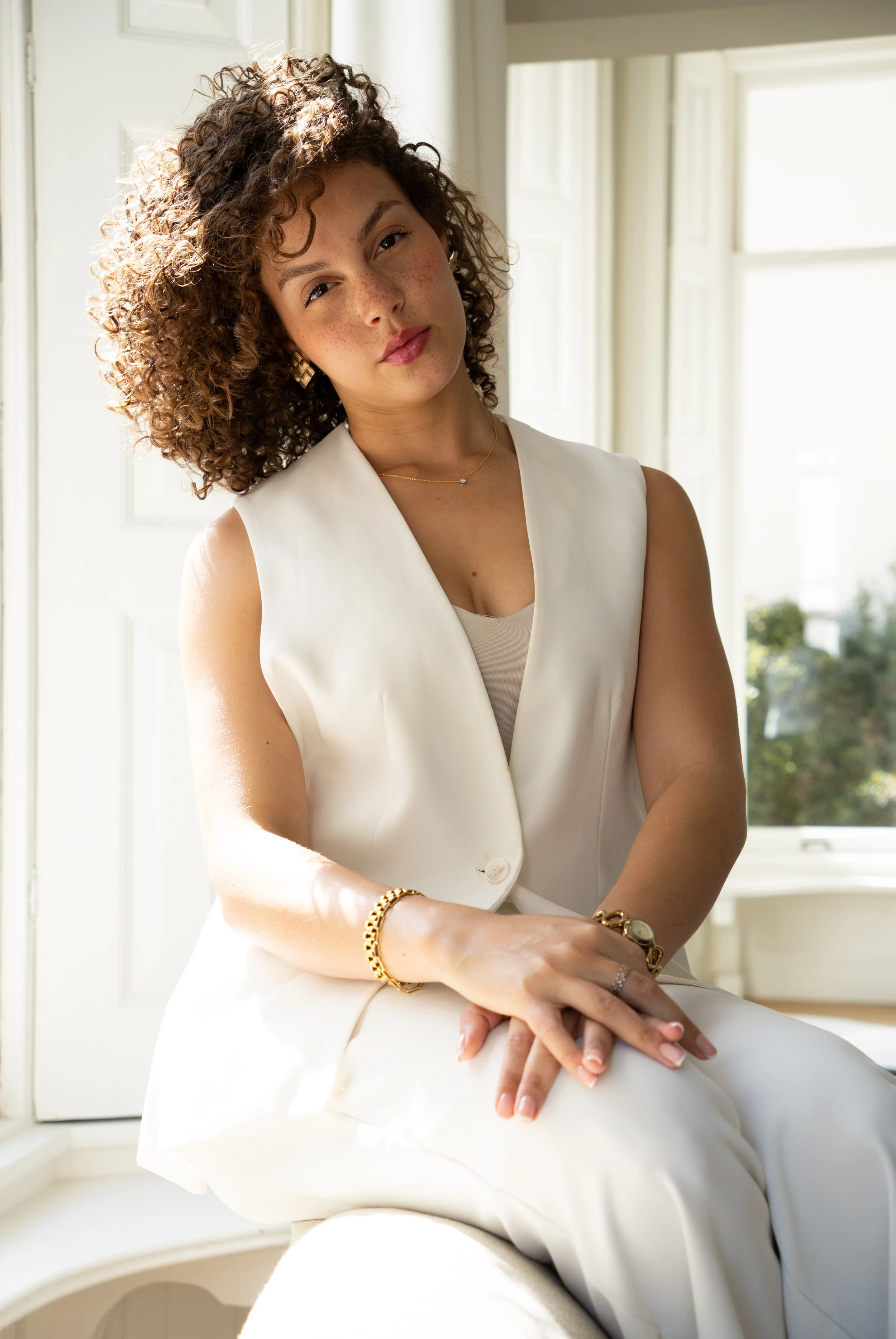 A woman with 3B and 3C curly brown hair and freckles sitting in front of a window, wearing a white sleeveless blazer, beige top, and gold jewelry, looking at the camera.