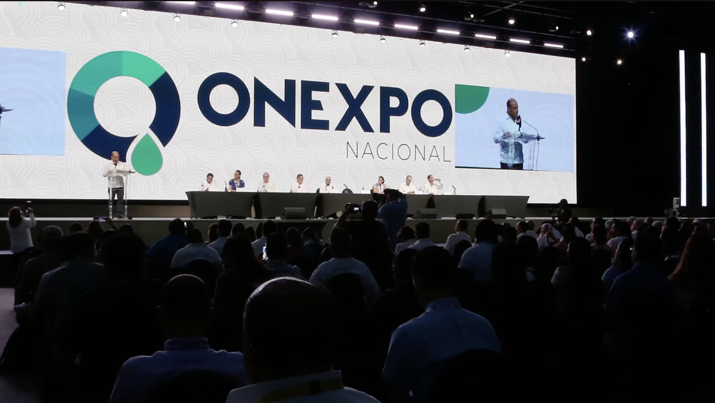 Escenario de conferencia con gran pantalla que lee 'ONEXPO NACIONAL' y varios asistentes en la audiencia.