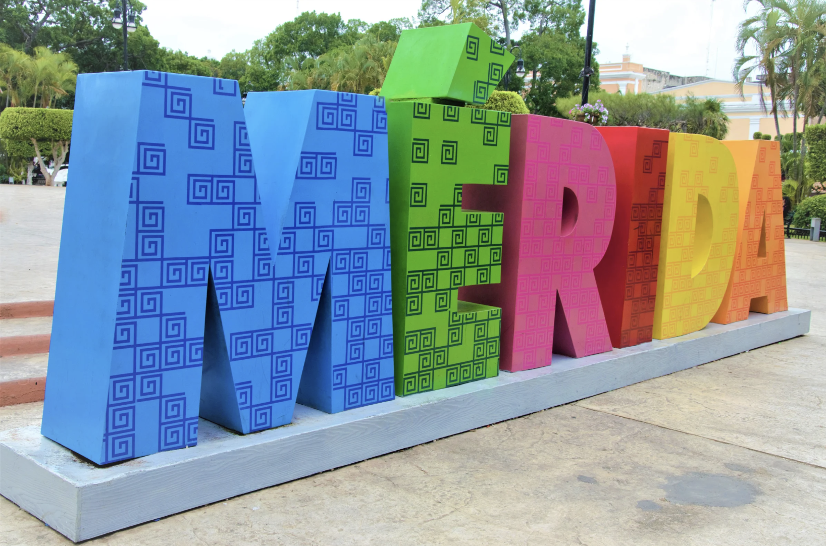 Letra grande y colorida que dice 'MERIDA' con fondo de árboles y palmeras.
